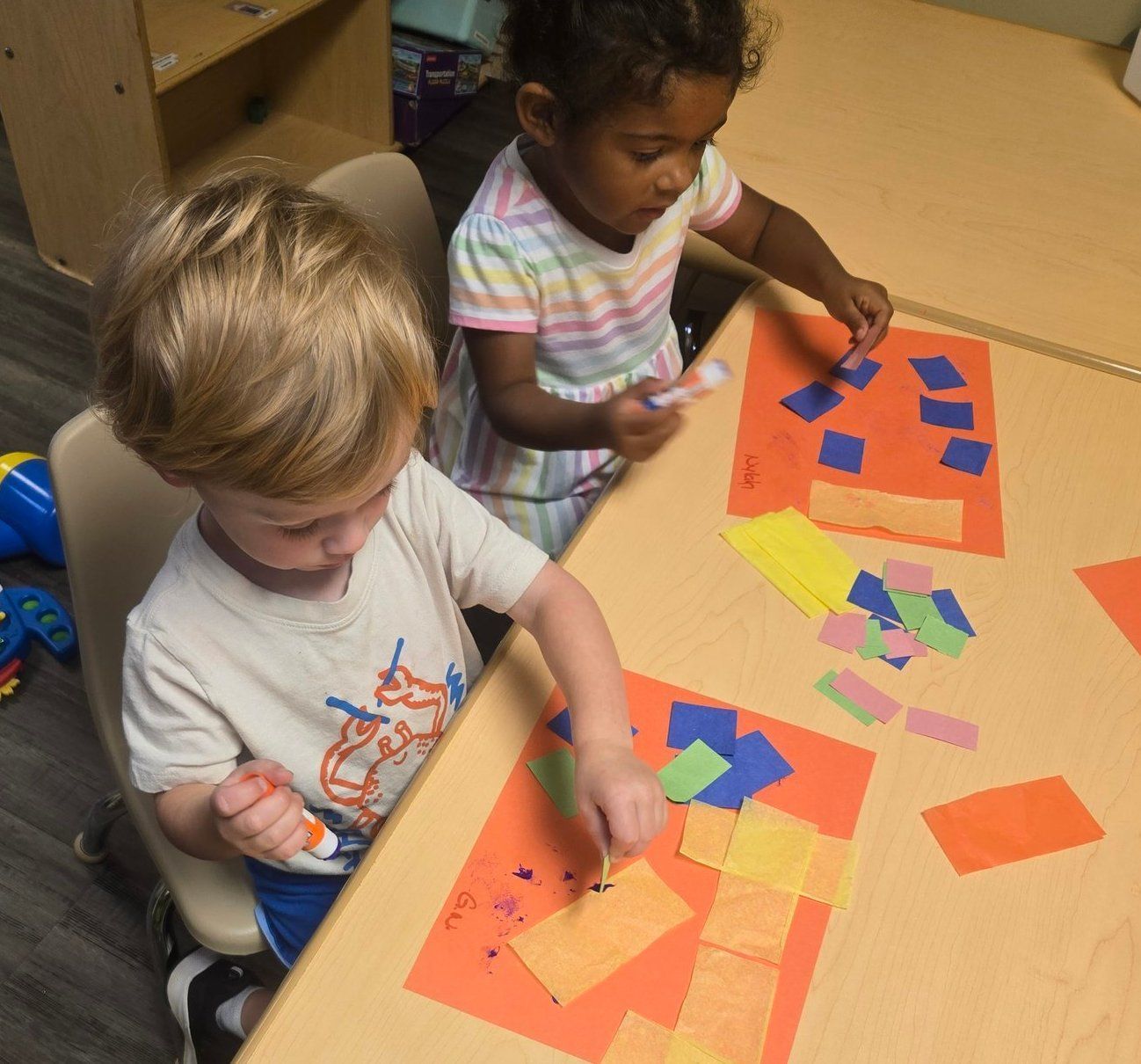 Two children at a table, gluing colorful paper pieces onto orange paper, indoors.