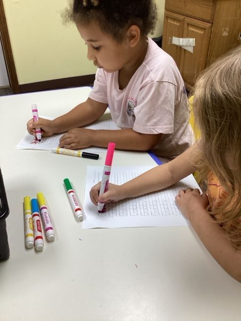 Two children seated at a table, using markers to draw on paper. Markers are scattered on the table.