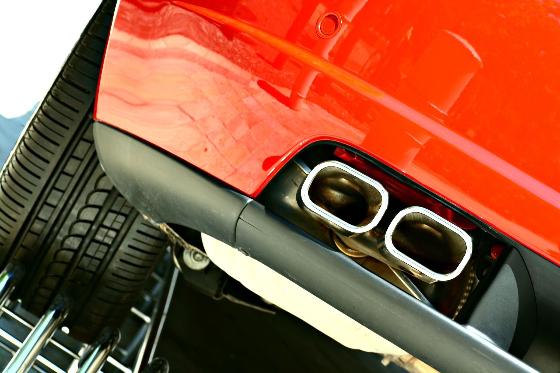 Red car's rear bumper with two chrome exhaust pipes and a black trim, and a partially visible tire.