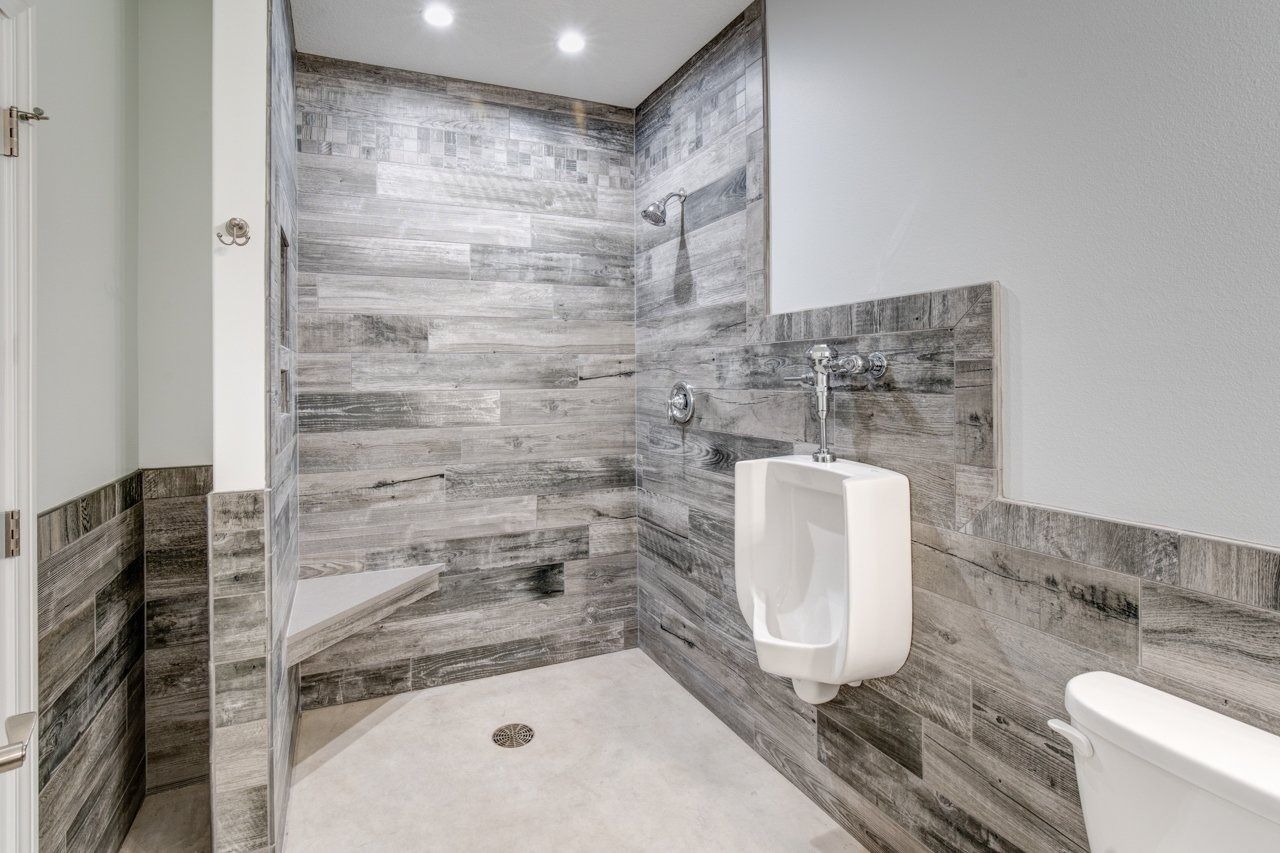 Walk-in shower with a urinal and toilet; gray wood-look tile, white fixtures, and recessed lighting.