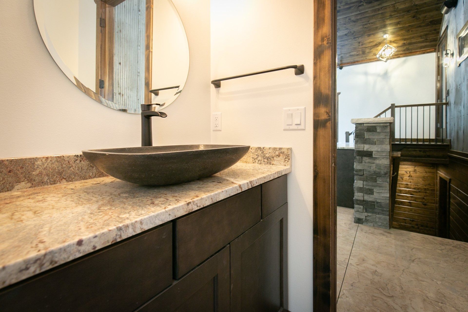 Bathroom with dark wood vanity, stone countertop, bowl sink, and round mirror.