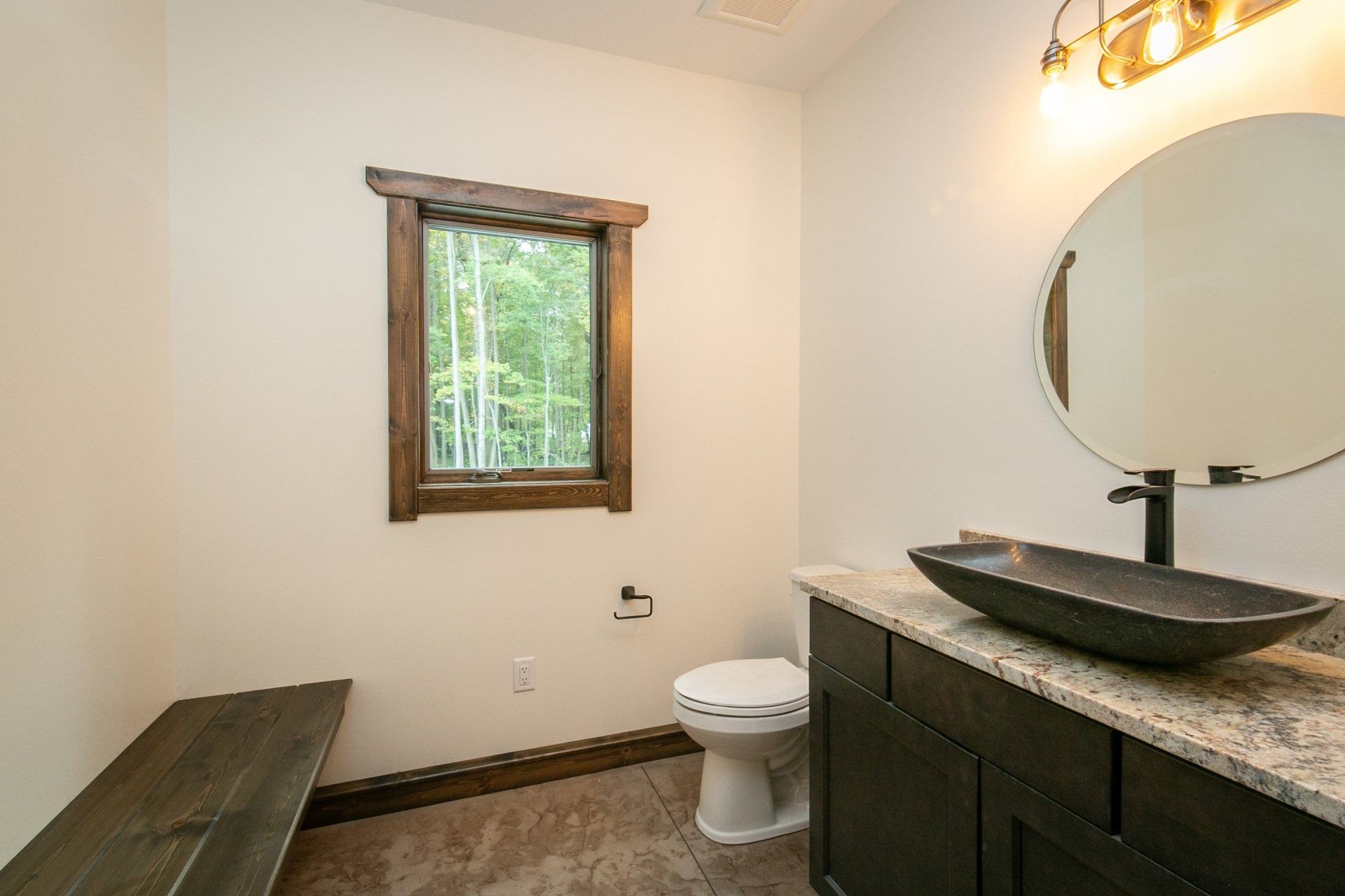 Bathroom with dark vanity, black sink, round mirror, window with wood trim, and a toilet.