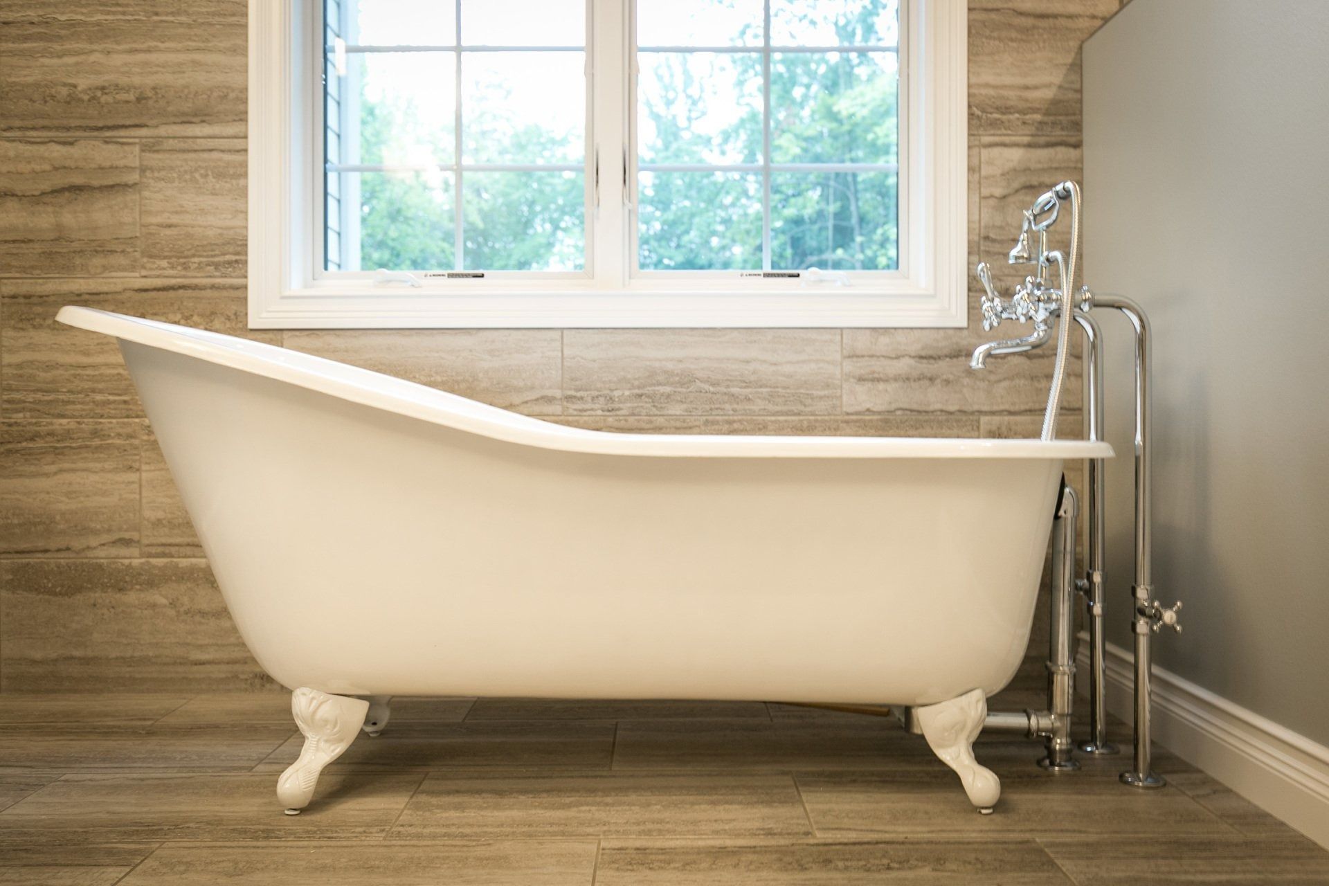 White clawfoot bathtub near a window, with a silver faucet fixture and light-colored wood-look tiled walls.