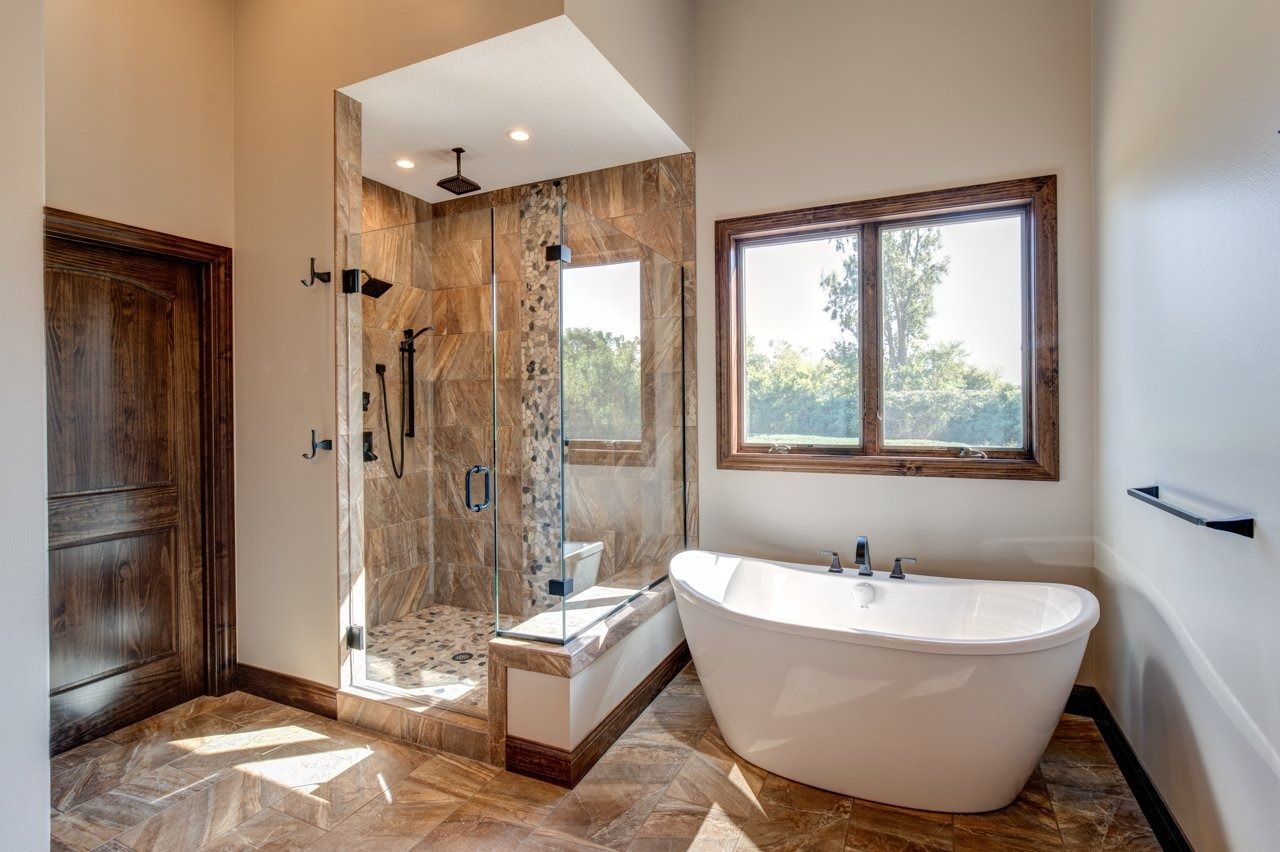 Spacious bathroom with glass shower, freestanding tub, wooden door and window frame, and stone-like tile.