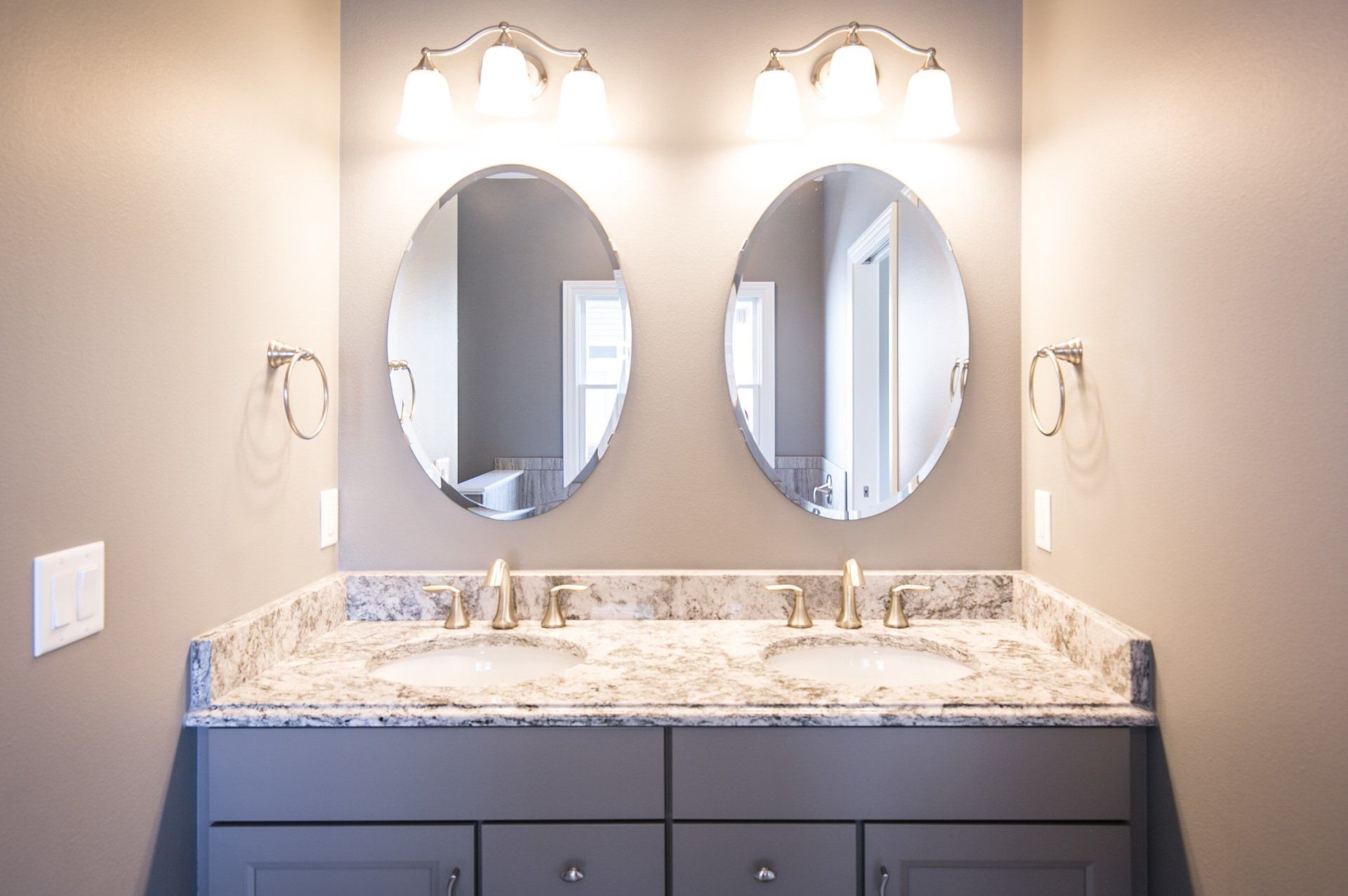 Bathroom vanity with two oval mirrors, two sinks, granite countertop, grey cabinets, and wall-mounted lighting.