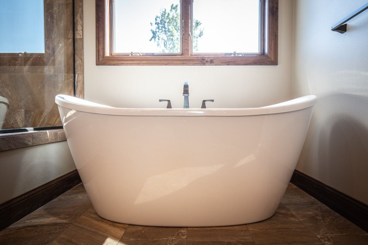 Freestanding white bathtub in bathroom, under a window with natural light.