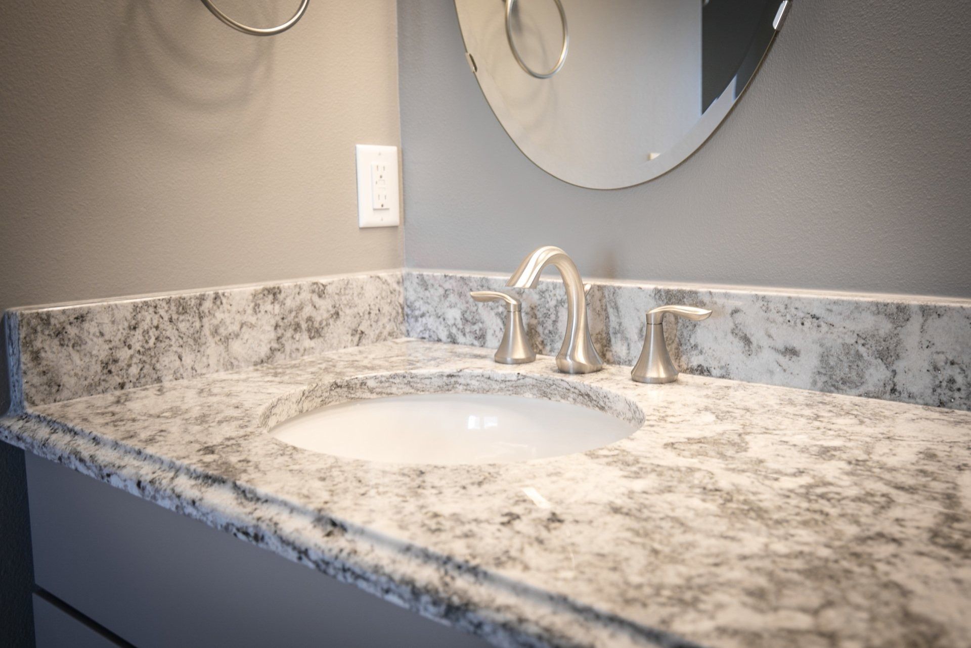 Bathroom with granite countertop, oval sink, silver faucet, round mirror, gray walls.