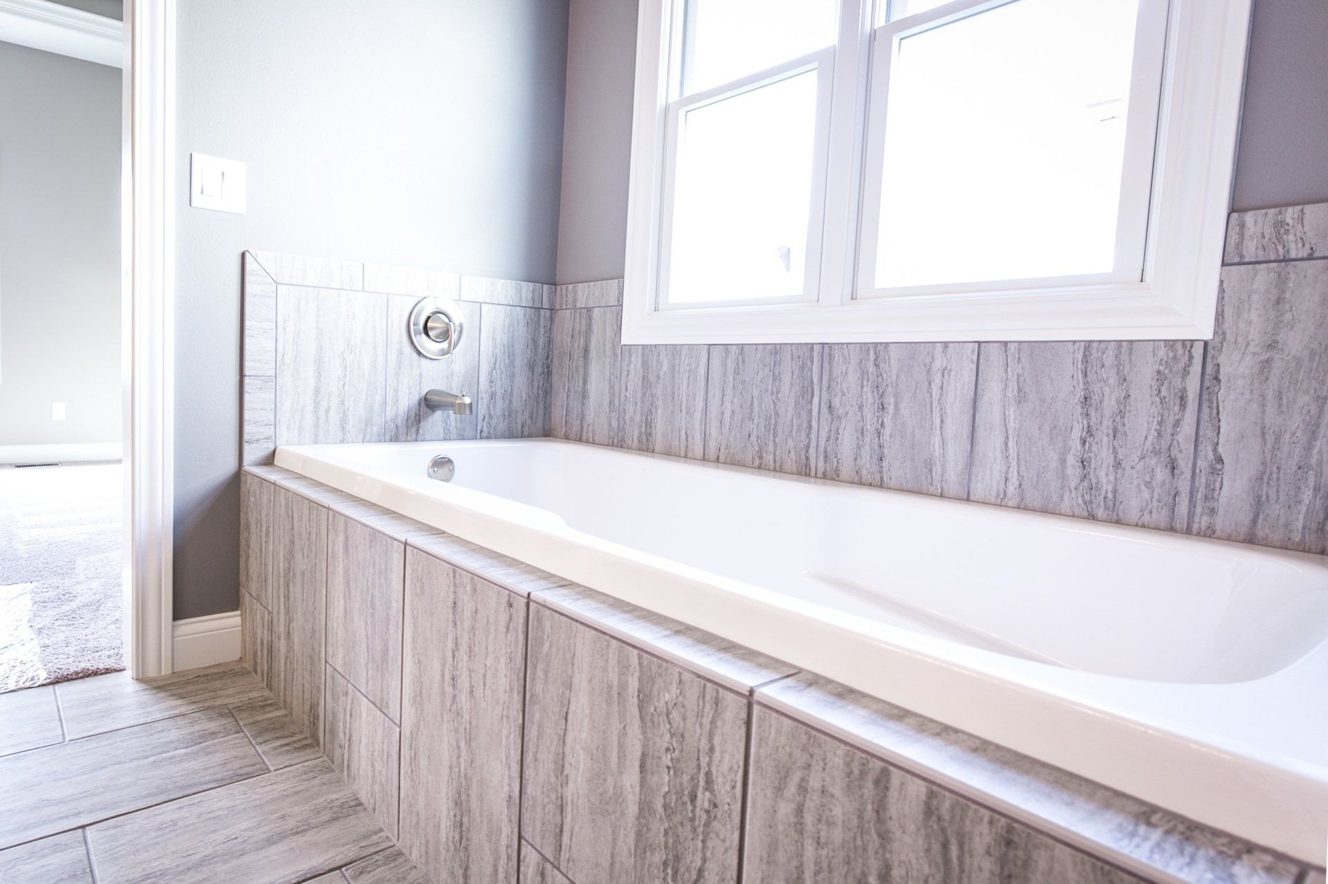 Modern bathroom with a white bathtub surrounded by gray tiled walls and a window.