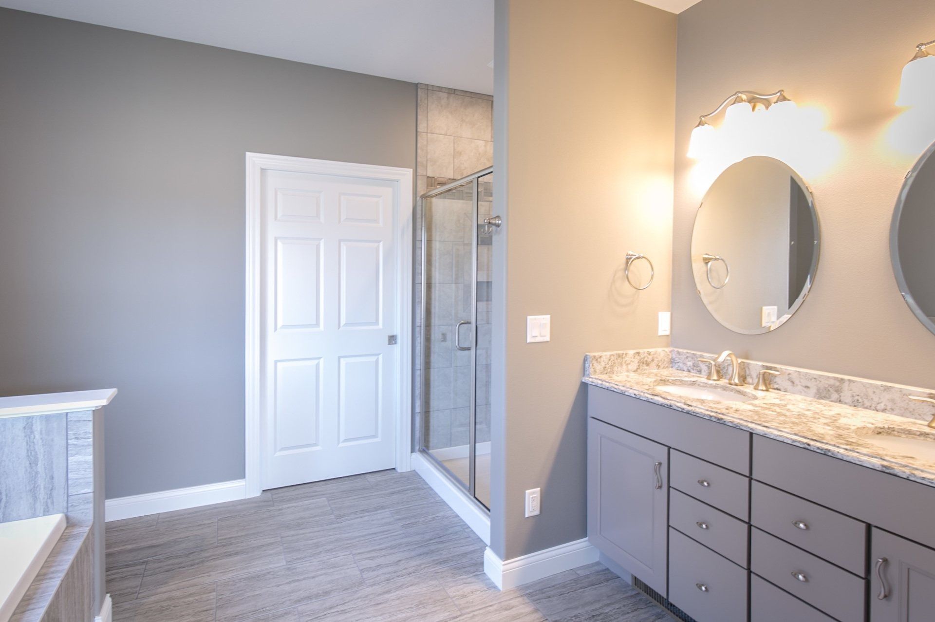 Gray bathroom with double vanity, oval mirrors, and a glass shower.