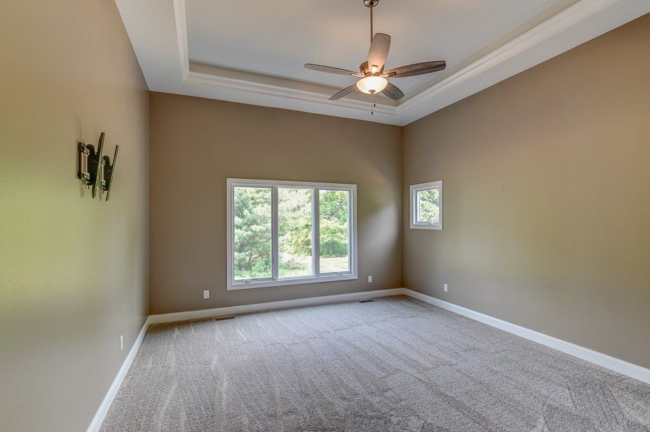 Empty bedroom with beige walls, gray carpet, large windows, and a ceiling fan.