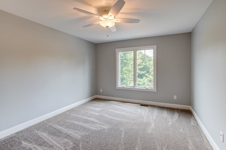 Empty bedroom with gray walls, beige carpet, window, and ceiling fan.