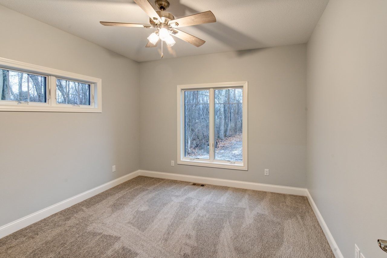 Empty bedroom with gray walls, two windows, and brown carpet. A ceiling fan is visible.
