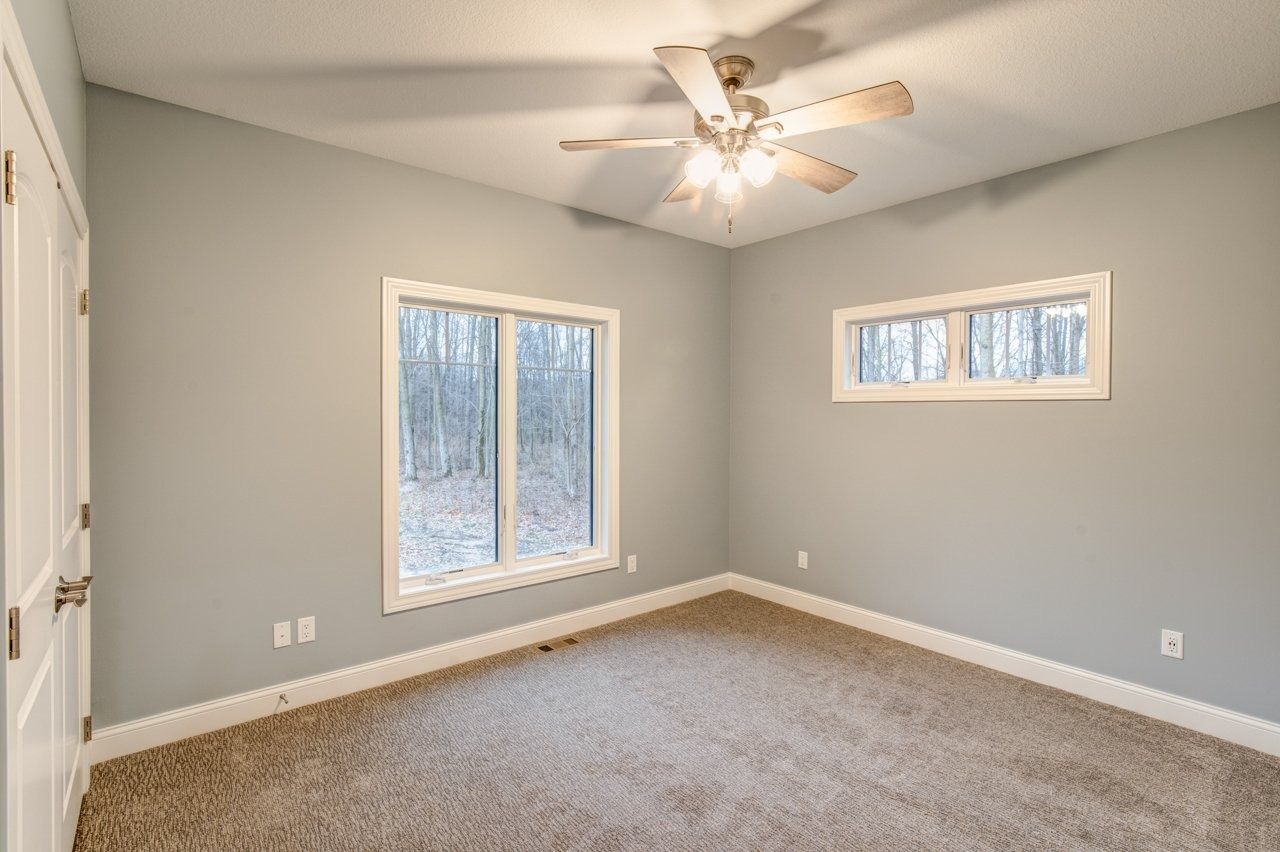 Empty bedroom with gray walls, two windows, tan carpet, and a ceiling fan.