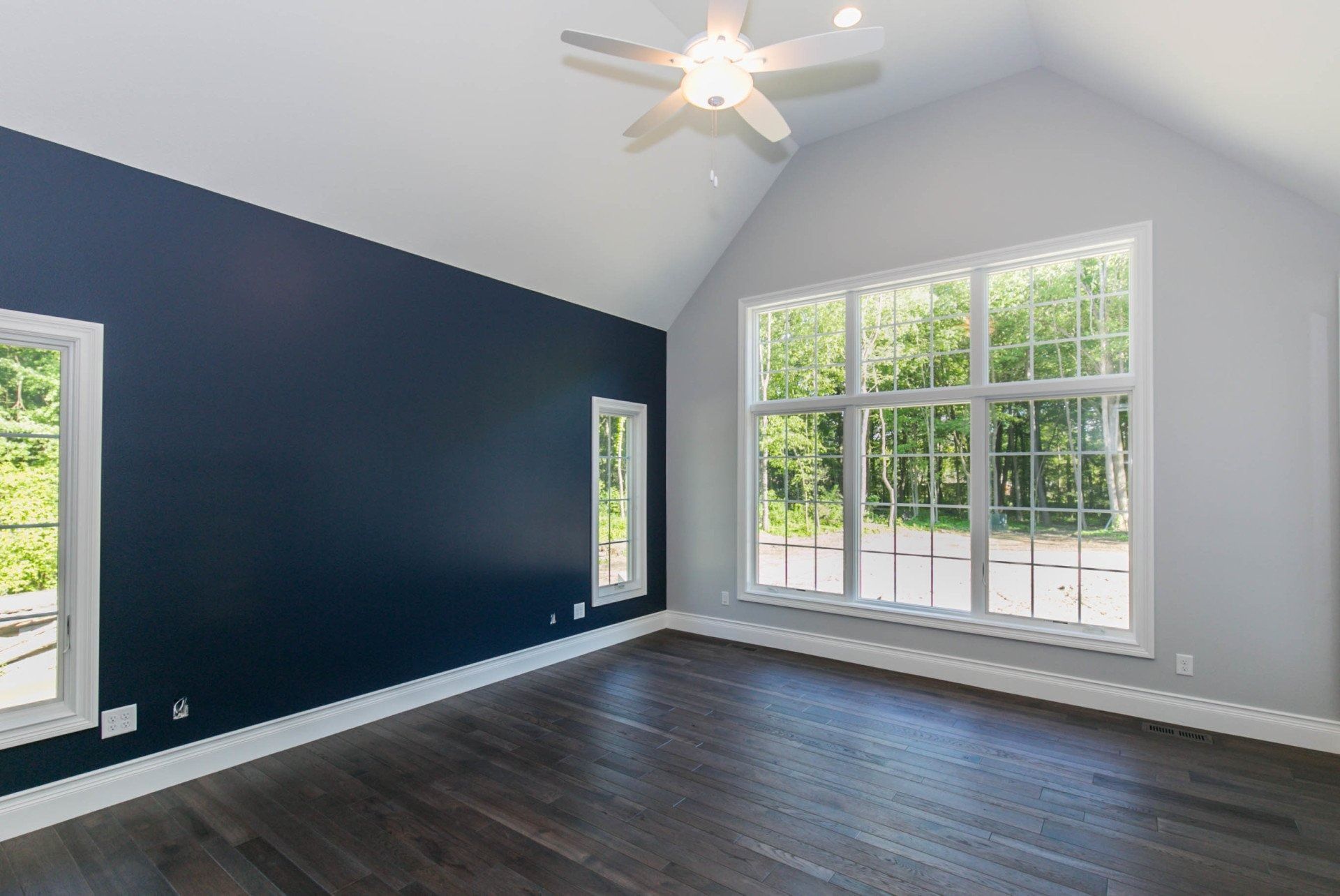 Empty bedroom with dark blue accent wall, white-framed windows, hardwood floor, and a ceiling fan.