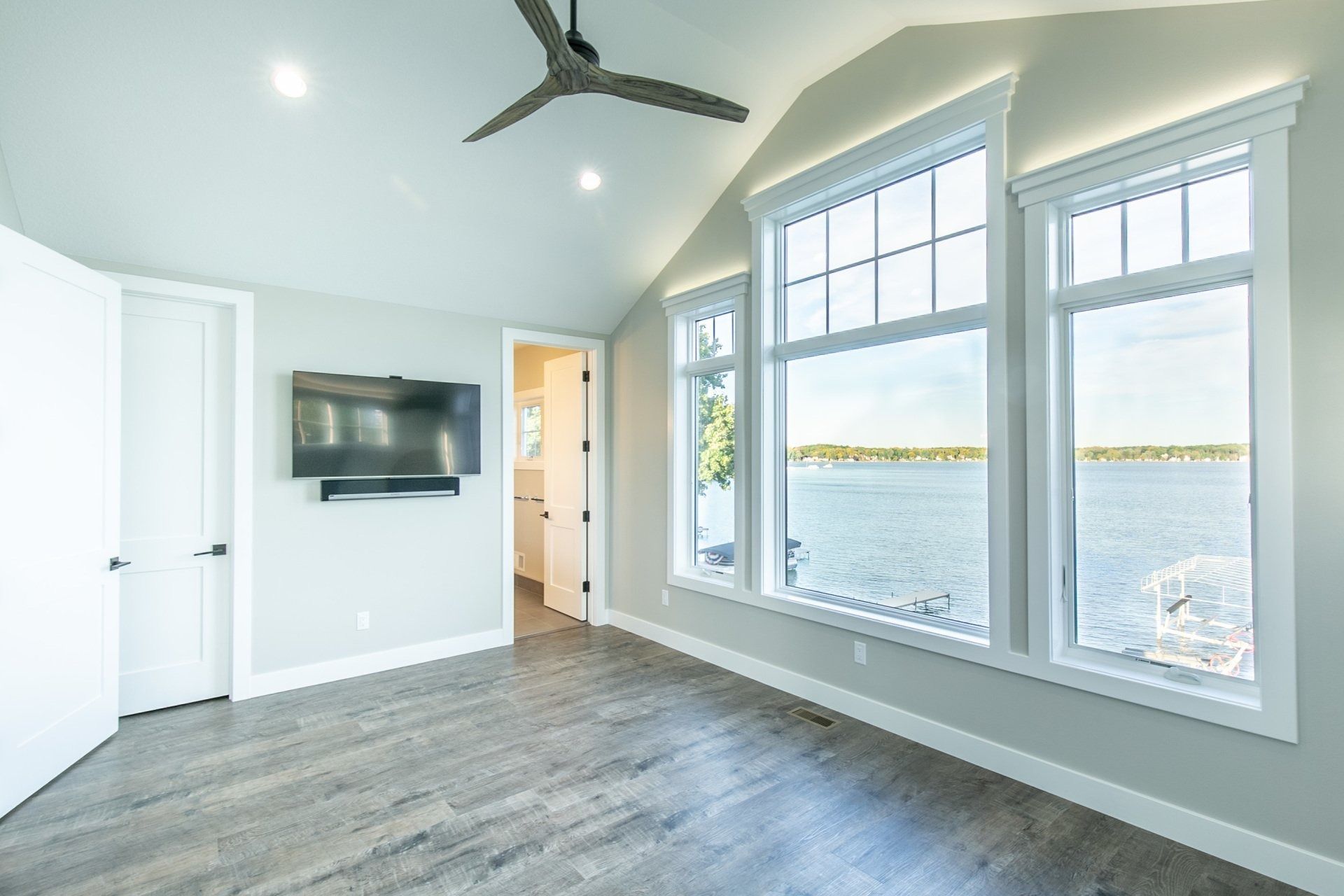 Spacious bedroom with lake view windows, a TV, and a door to a bathroom. Gray walls, wood-look flooring.