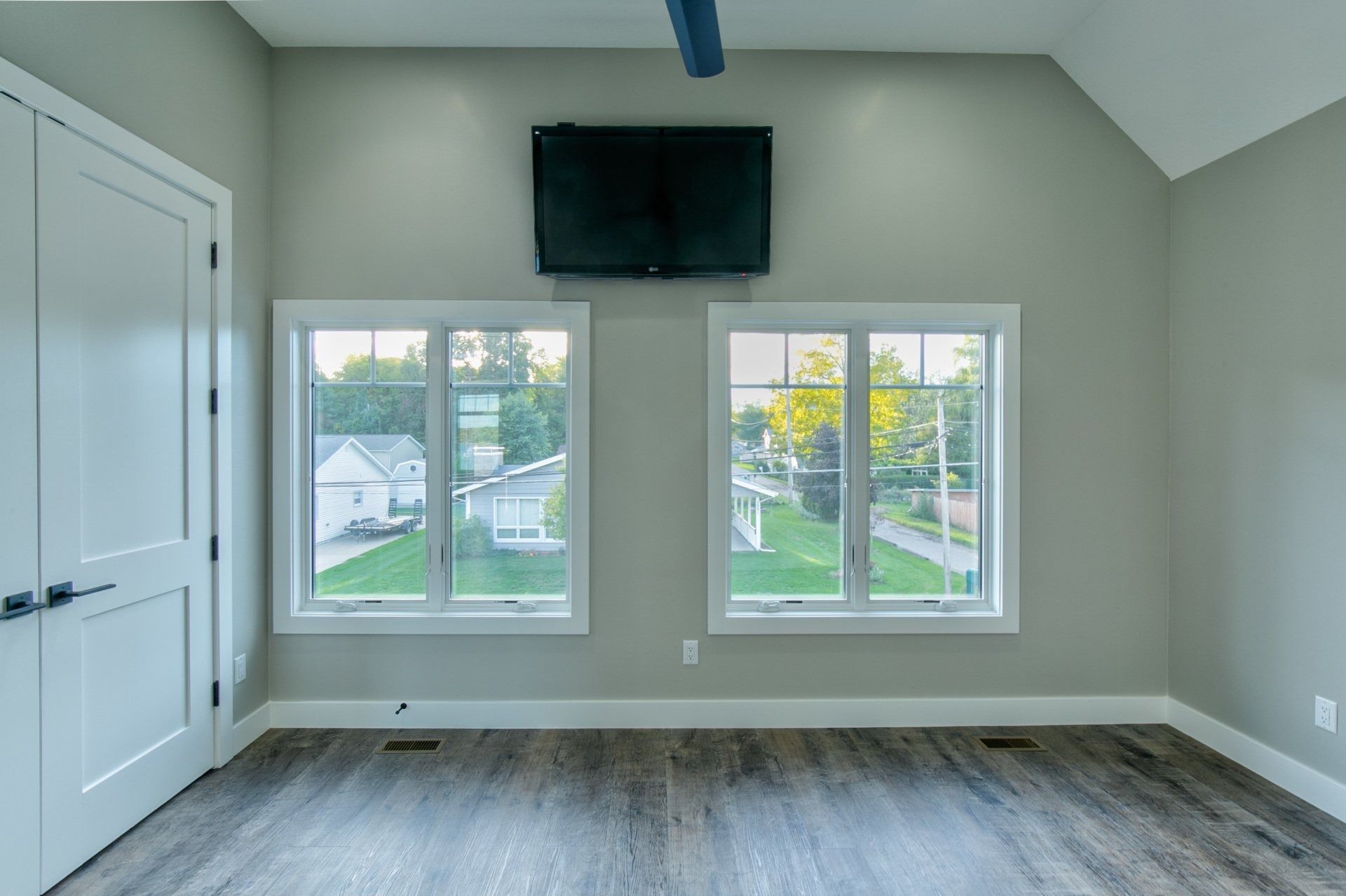 Empty room with two windows, a mounted TV, white door, and gray walls.