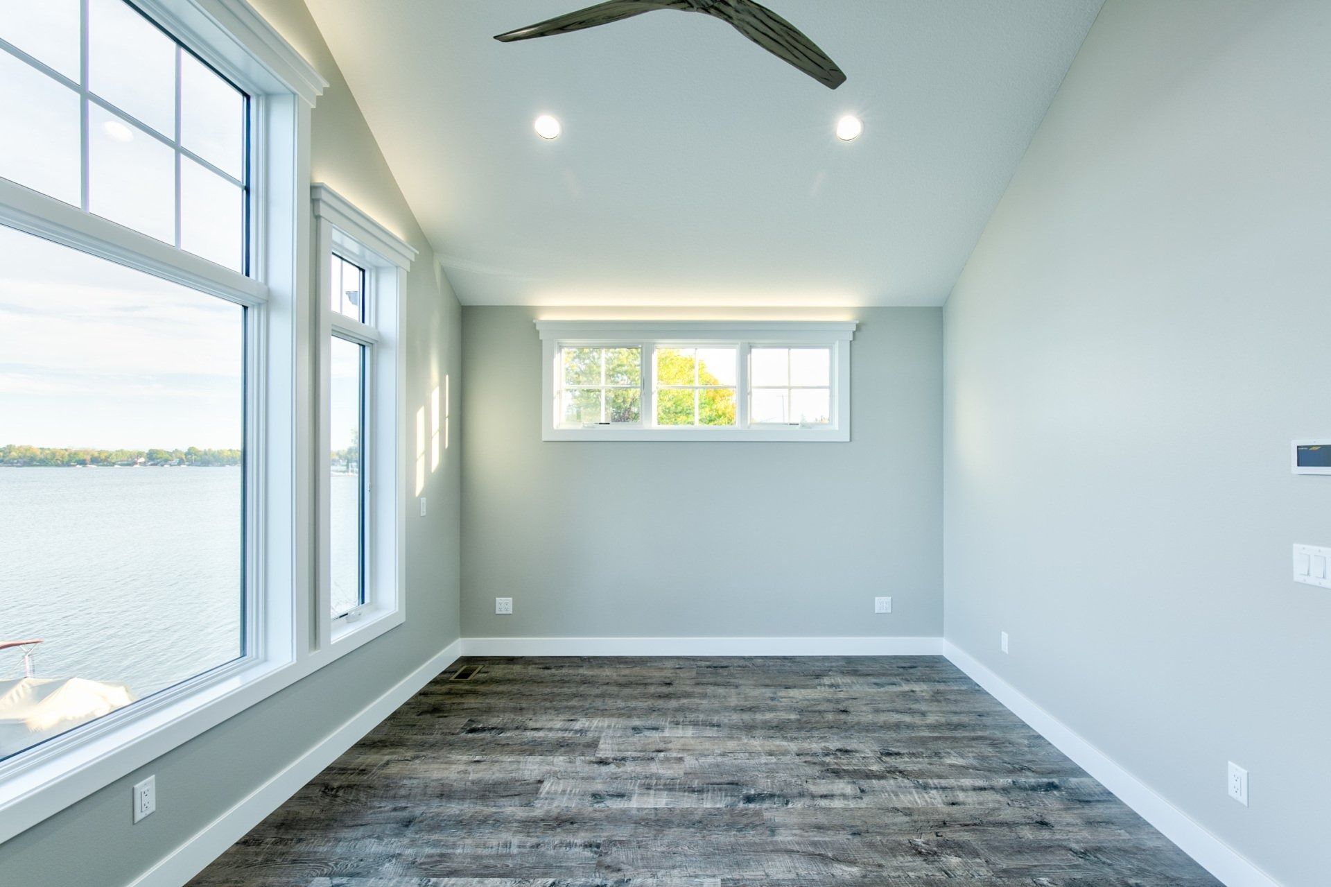 Empty room with large windows overlooking water, gray walls, dark wood-look floor, and white trim.