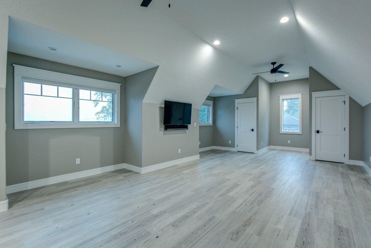 Spacious, neutral-toned room with angled ceilings, light wood floors, two windows, and mounted television.
