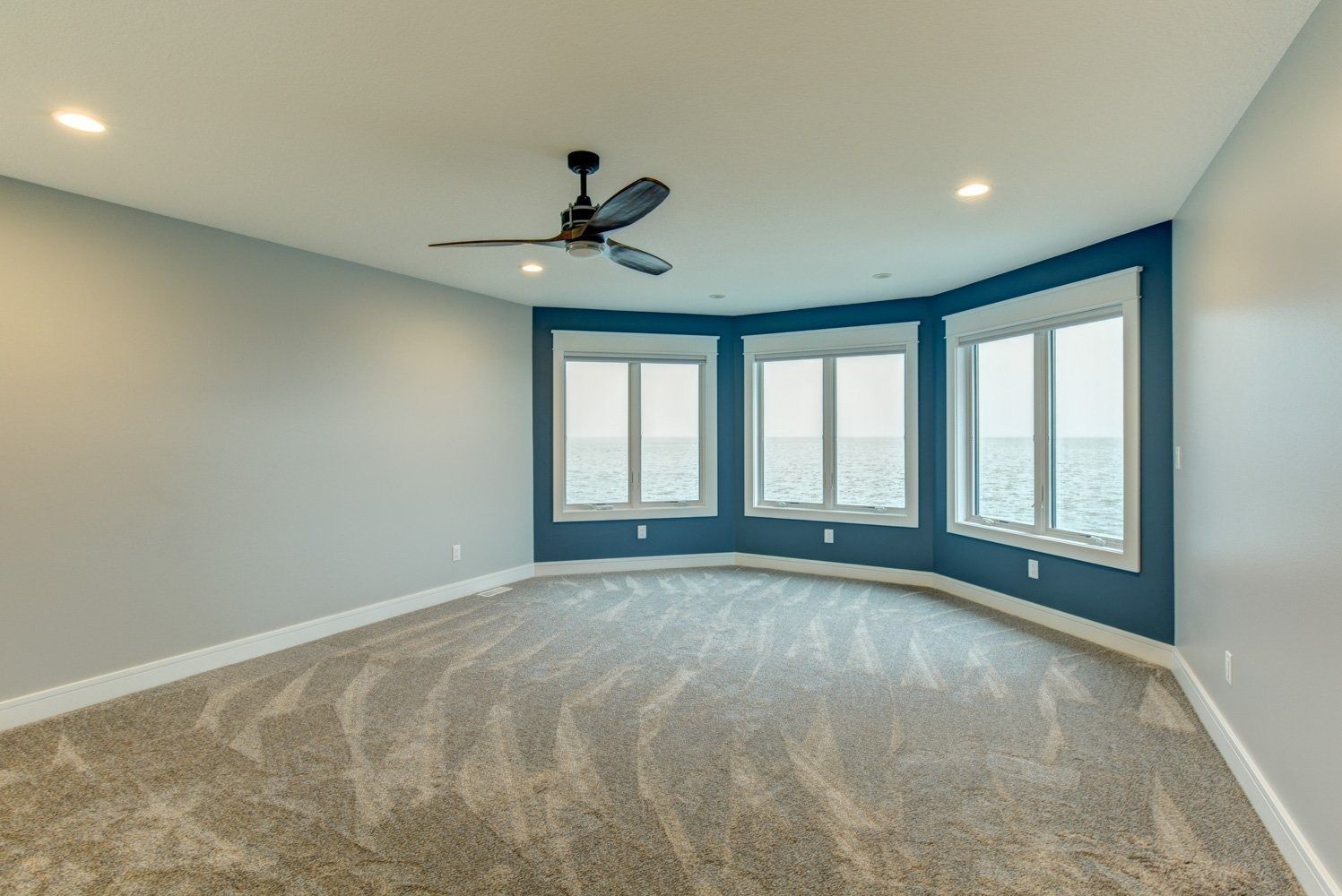 Empty room with gray carpet, blue accent wall, and three large windows.