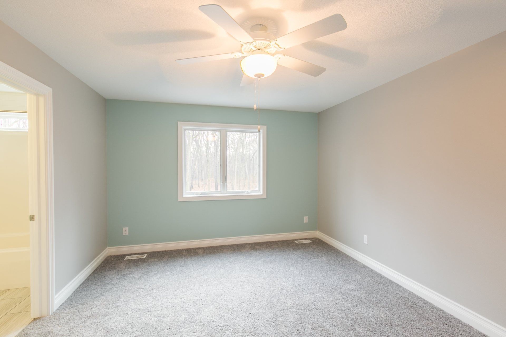 Empty bedroom with gray carpet, light blue wall, window, and ceiling fan.