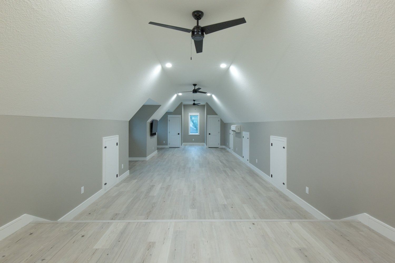 Spacious, light-filled attic room with gray walls, white trim, and light wood-look flooring. Two ceiling fans.