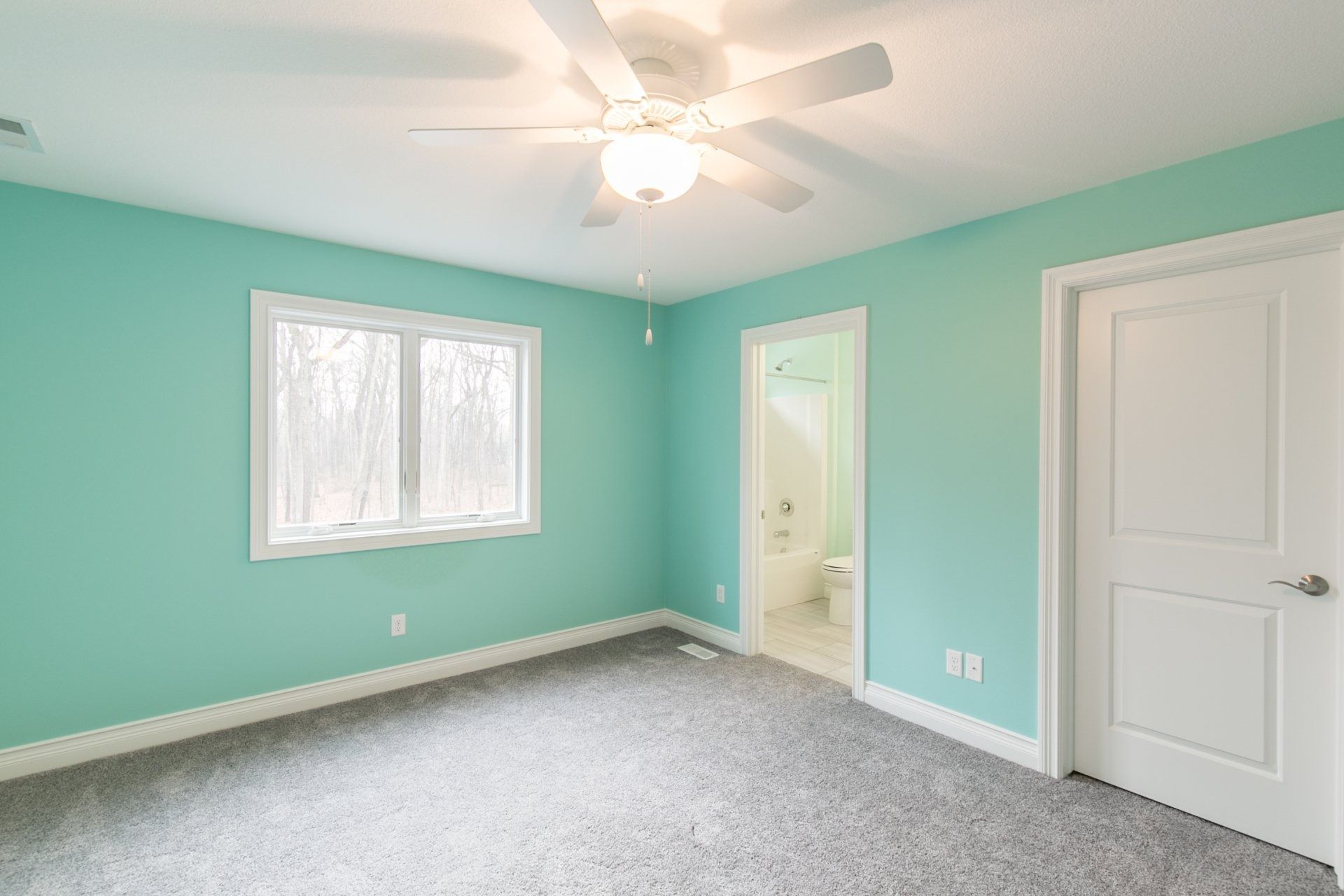 Empty bedroom with aqua walls, white door and trim, window, ceiling fan, and gray carpet.