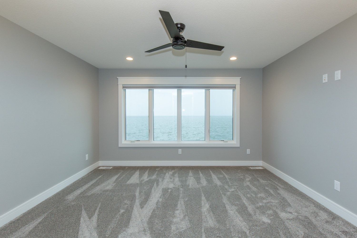 Empty room with large window overlooking water. Gray walls, carpet, ceiling fan.