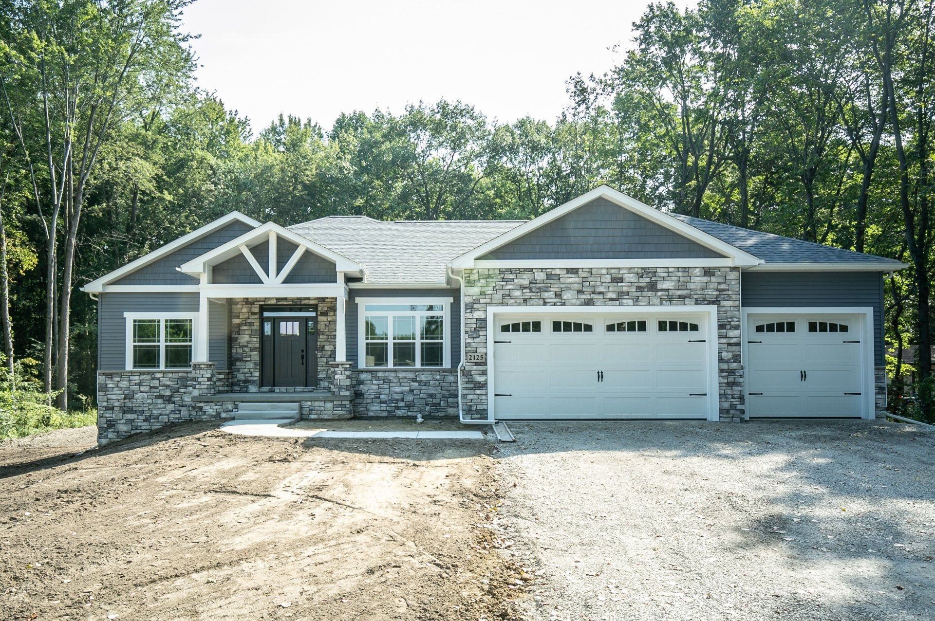 New, single-story house with stone and gray siding, three-car garage, and gravel driveway, surrounded by trees.