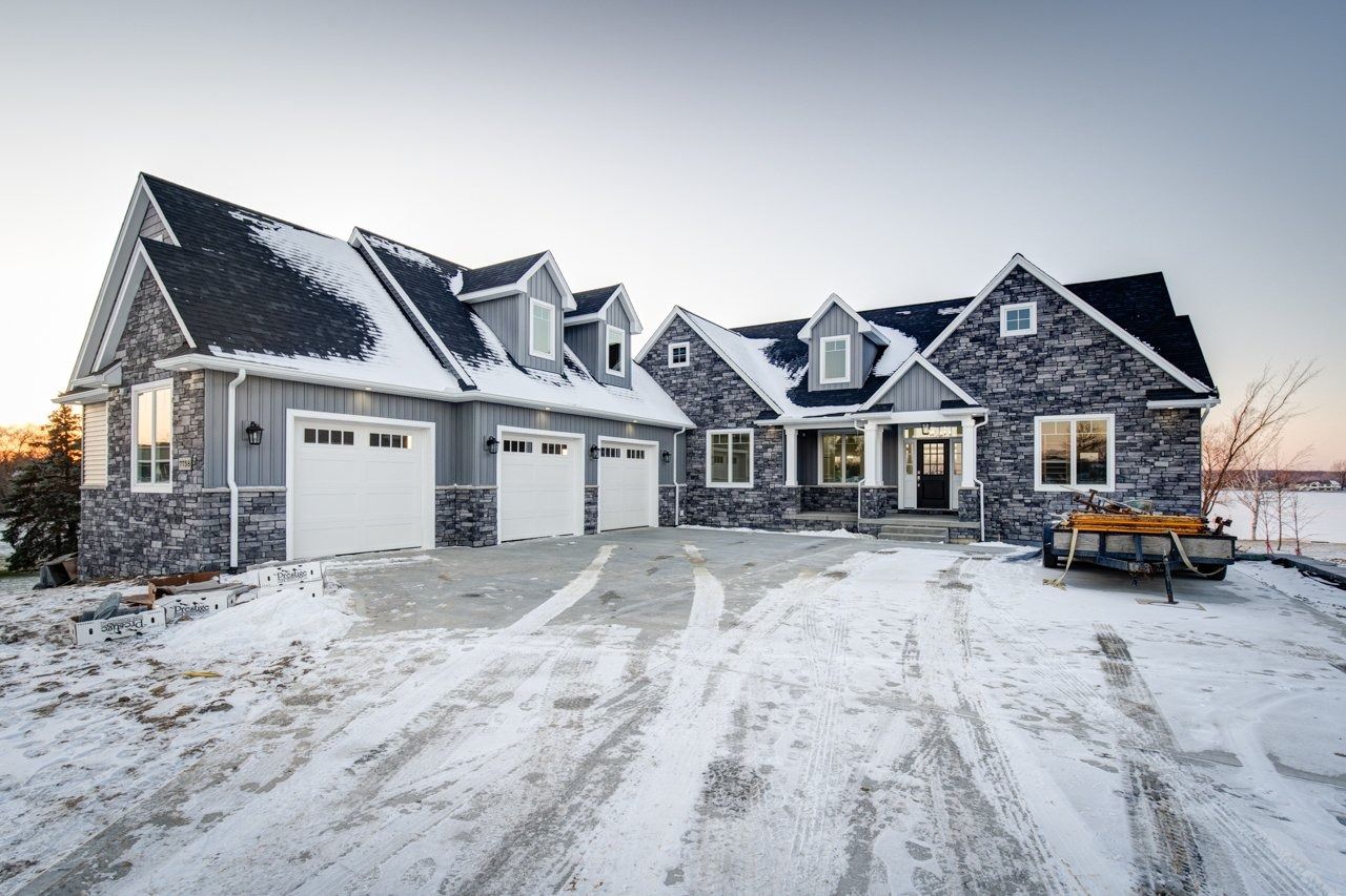 Snow-covered modern house with gray stone and siding, three-car garage, and a snow removal tool.