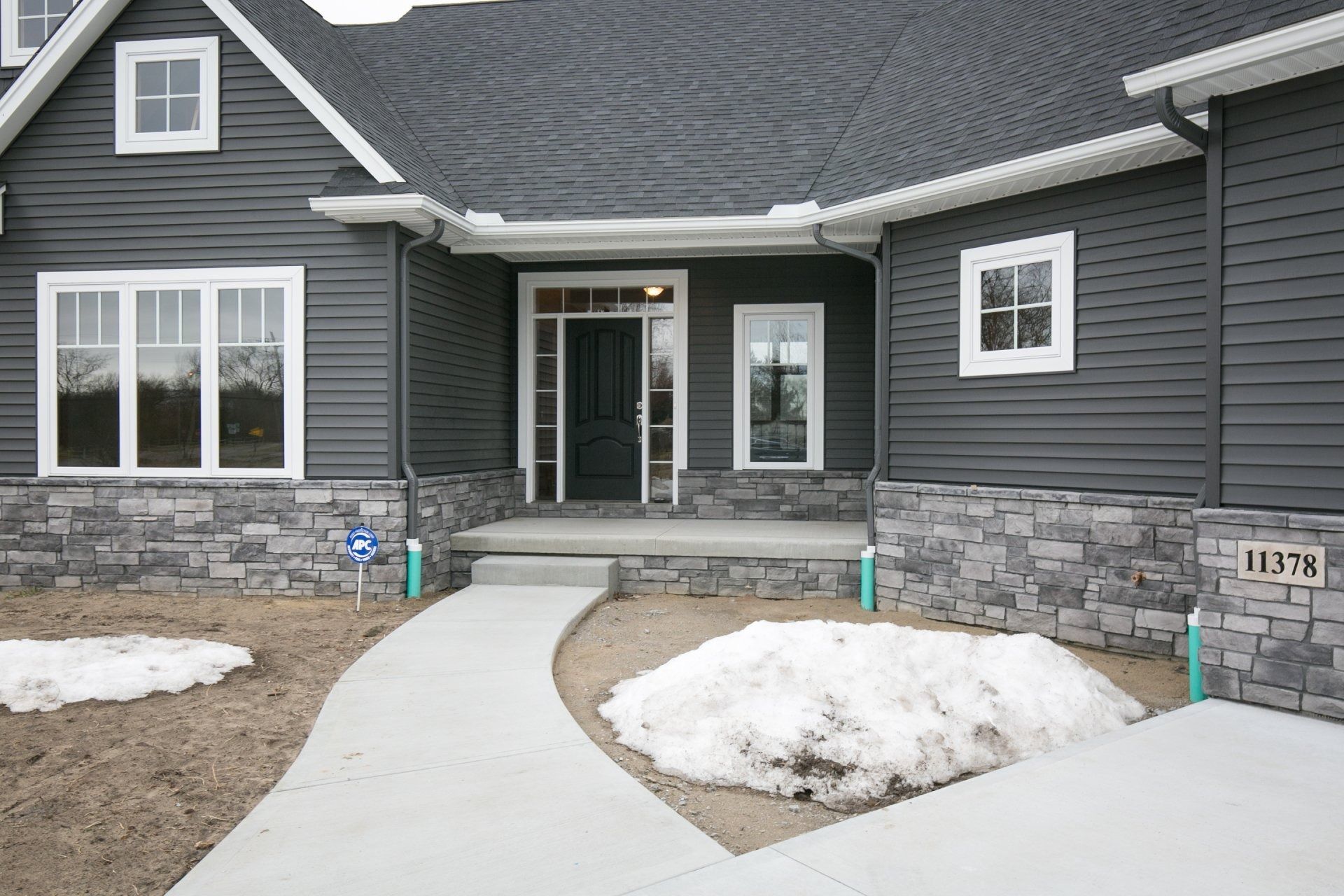 Gray house with white trim, stone base, and a curved concrete walkway with patches of snow.