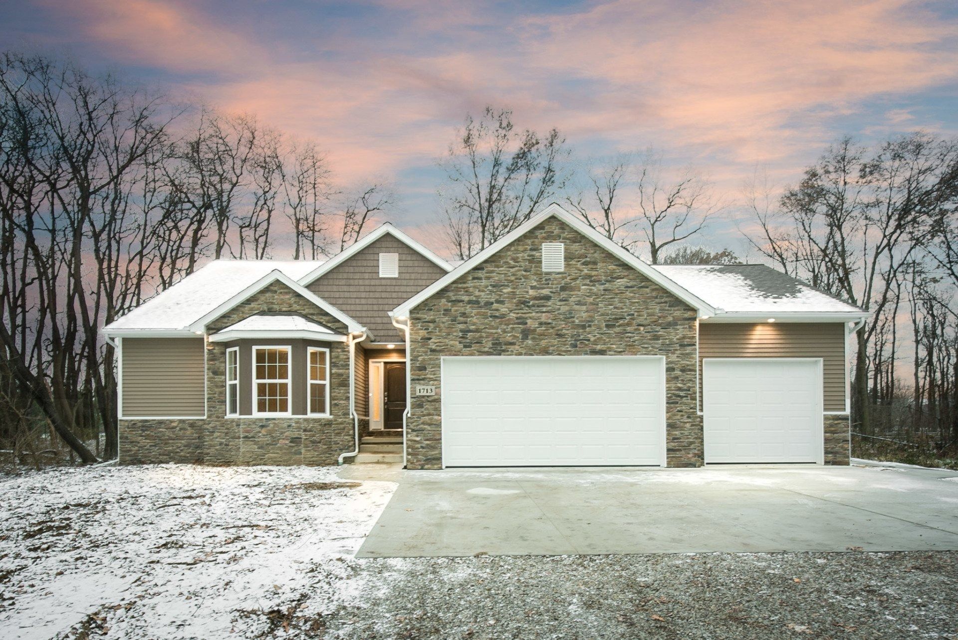 House exterior in winter; snow-covered ground, stone and siding facade, two-car garage, and cloudy sky.