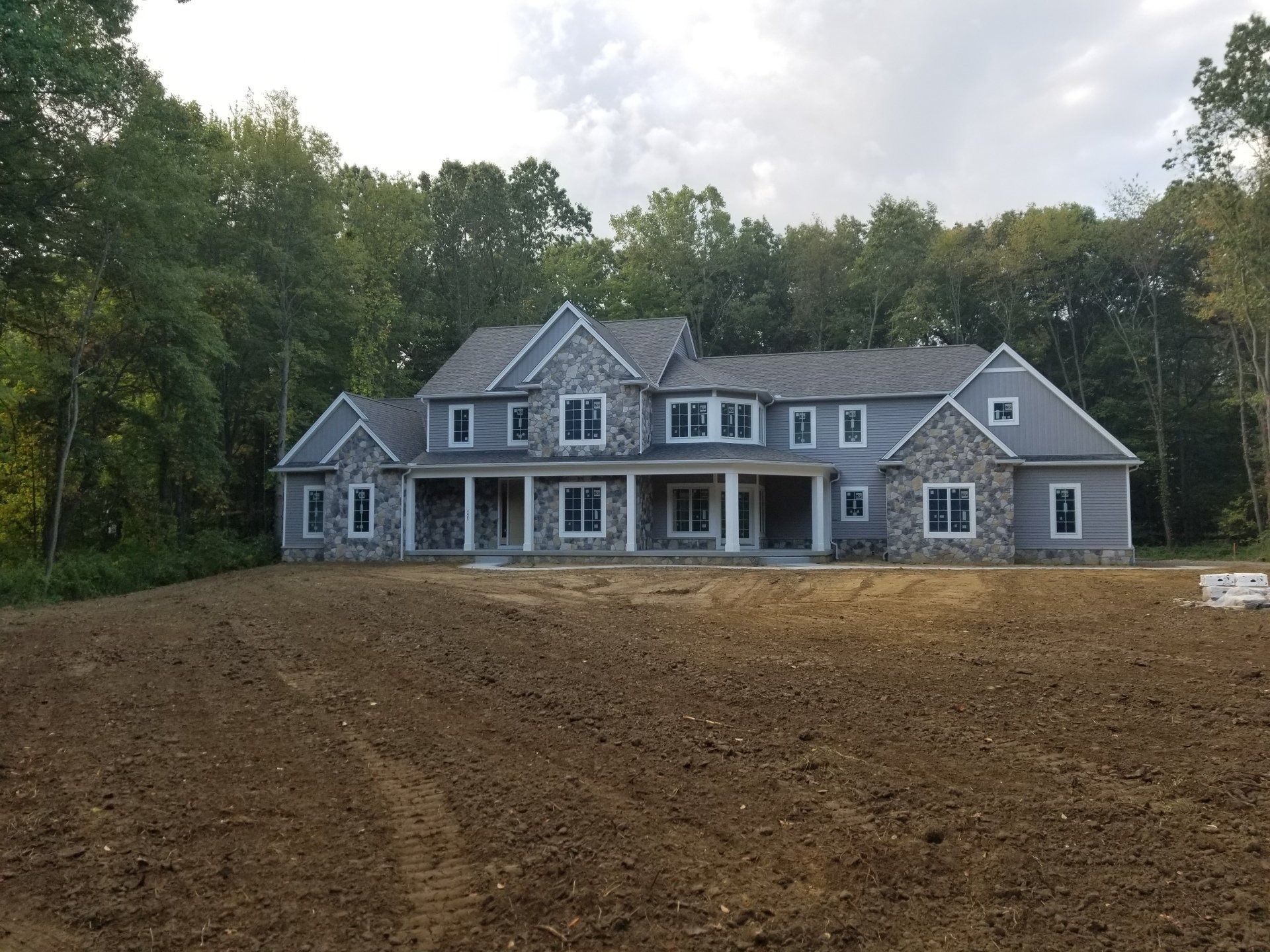 A large, two-story house with gray siding and stone accents, set in front of a freshly graded lawn with trees in the background.
