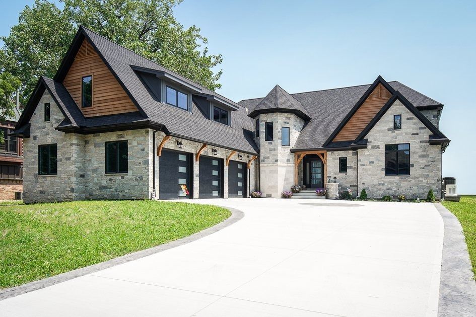 A modern stone house with a curved driveway, three black garage doors, and a turret.
