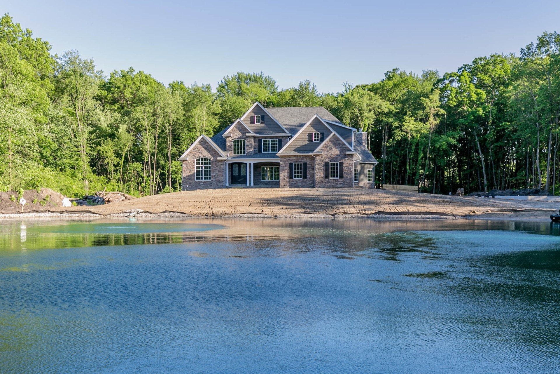 Large house with stone facade, built on a rise overlooking a lake, surrounded by trees.