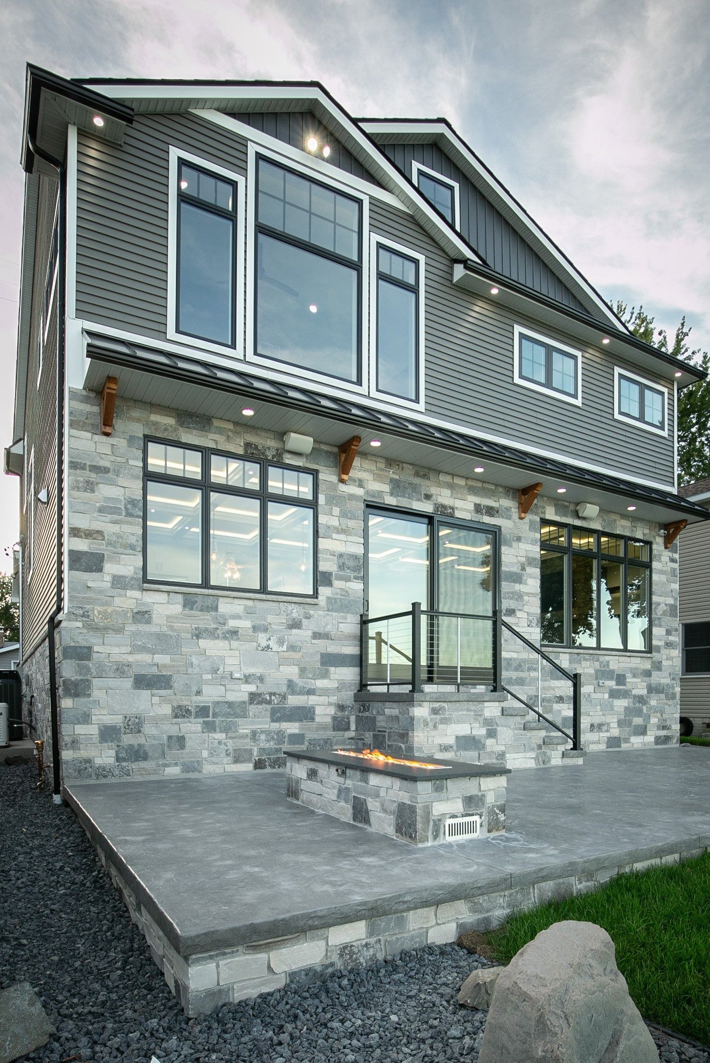 Gray stone and siding house with multiple large windows, dark trim, and a patio.