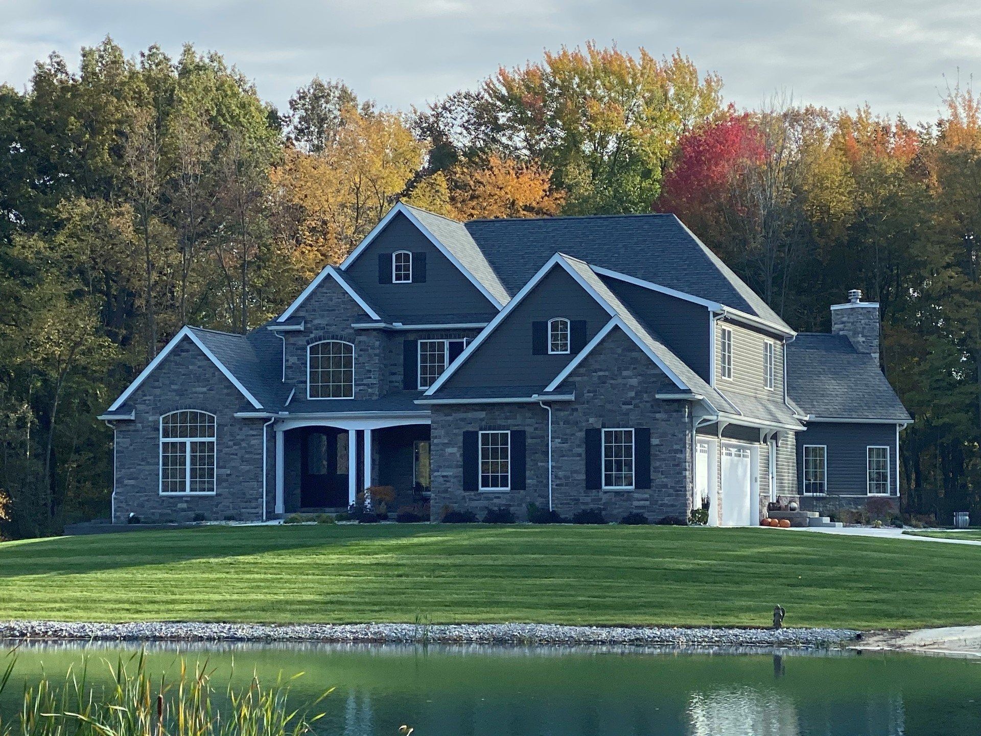 Two-story gray stone house with dark roof and green lawn beside a pond, fall foliage in the background.