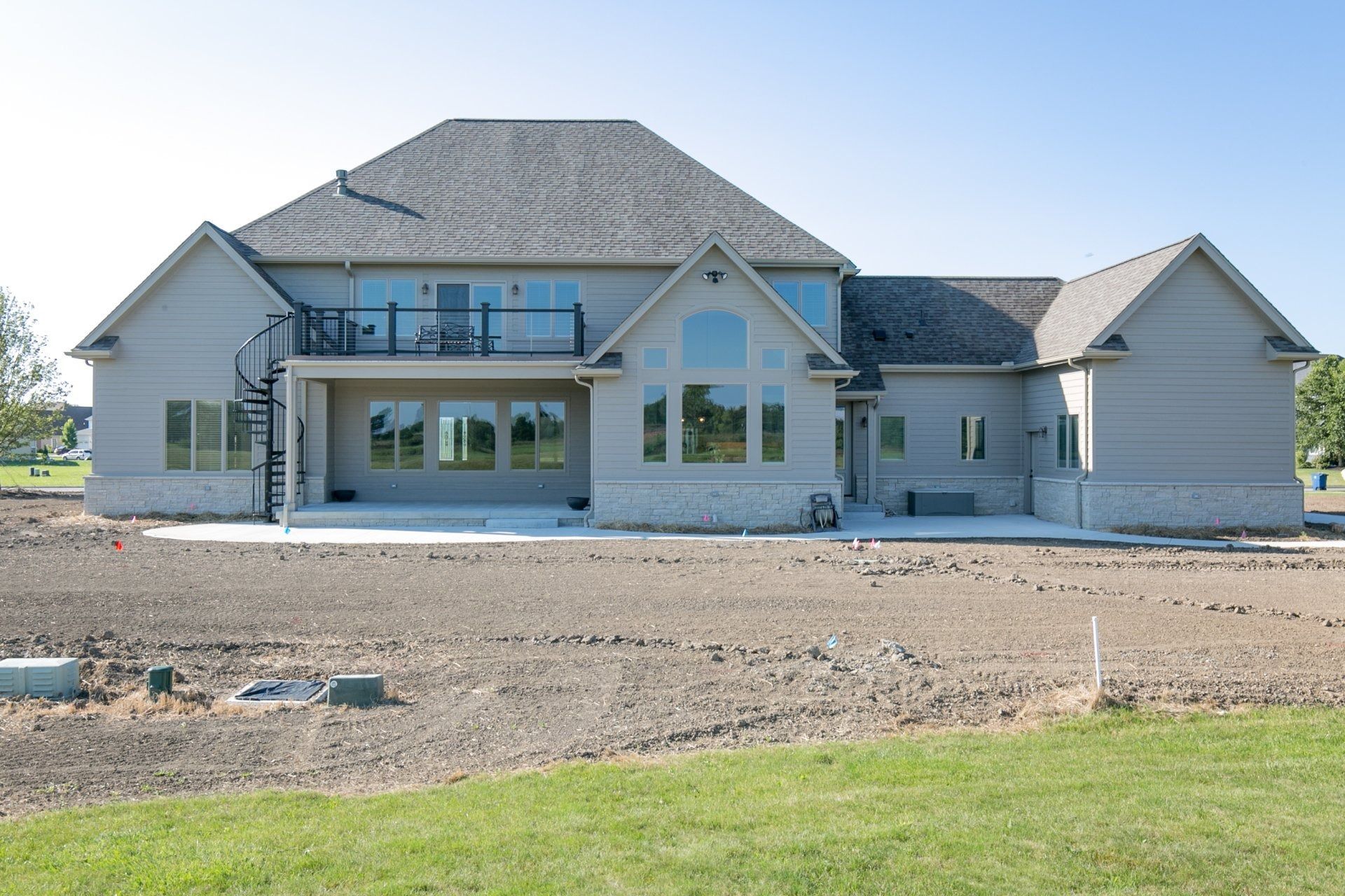 Rear view of a light gray house with a stone facade, gray roof, and large windows. Construction site with spiral staircase.