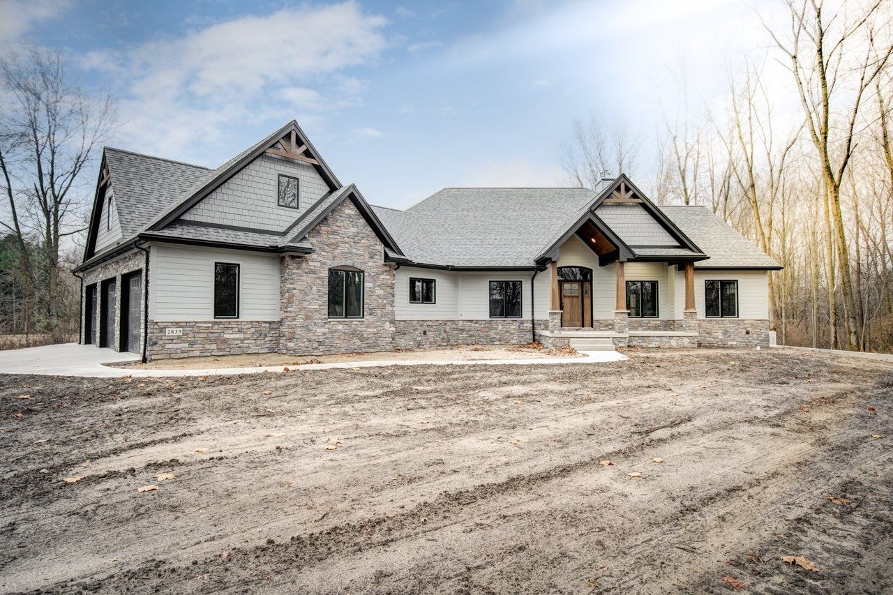 A new house under construction; gray stone and wood exterior with a garage, on a dirt lot, against a cloudy sky.