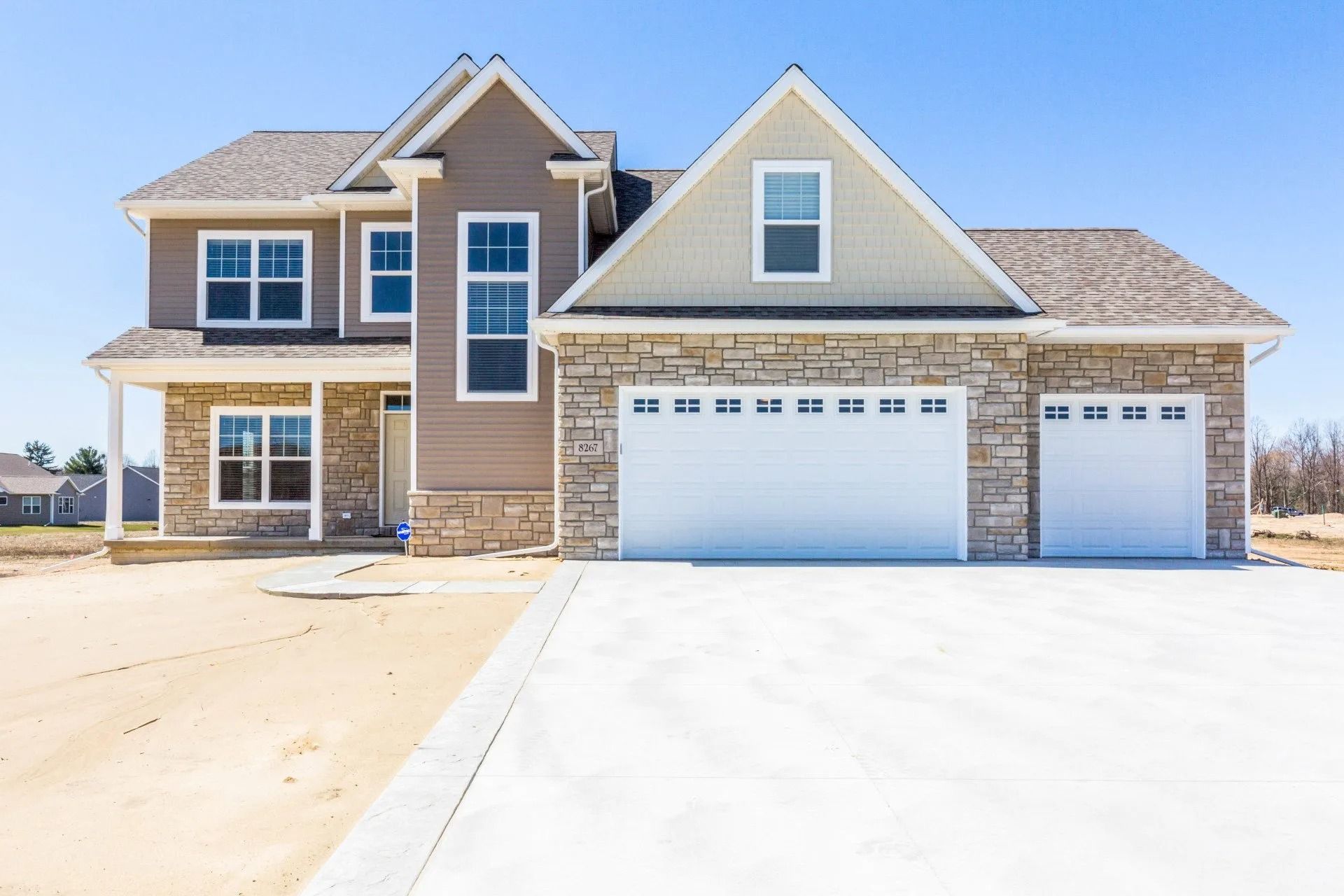 Two-story house with a stone and beige exterior, white garage doors, and a concrete driveway under a clear blue sky.