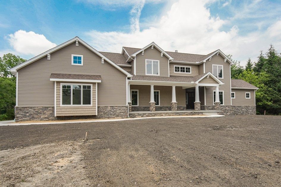 Two-story beige house with stone accents, columns, large windows, and unfinished gravel yard under a blue sky.