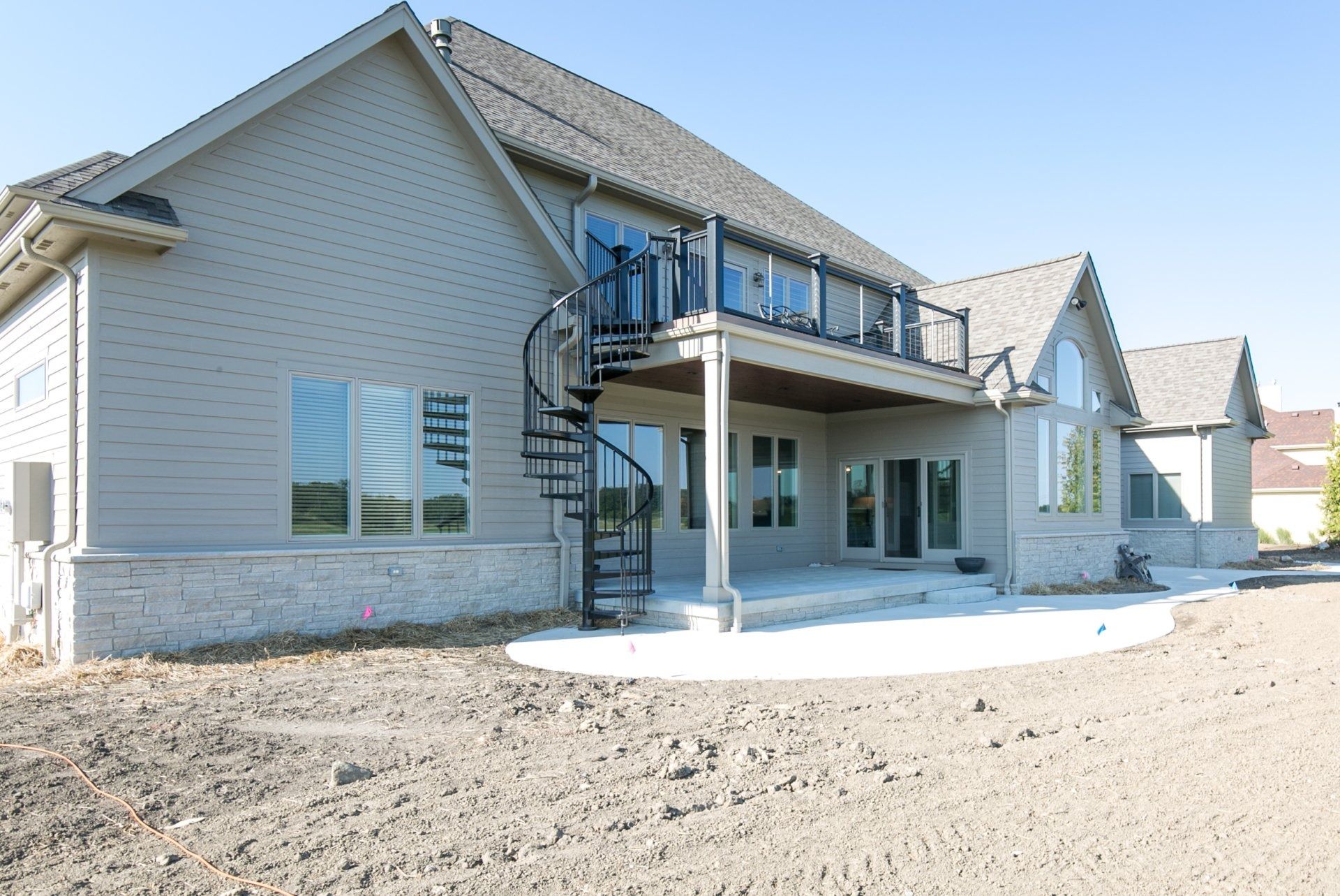 Back of a two-story beige house with a spiral staircase, deck, and patio. Gray roof and windows.