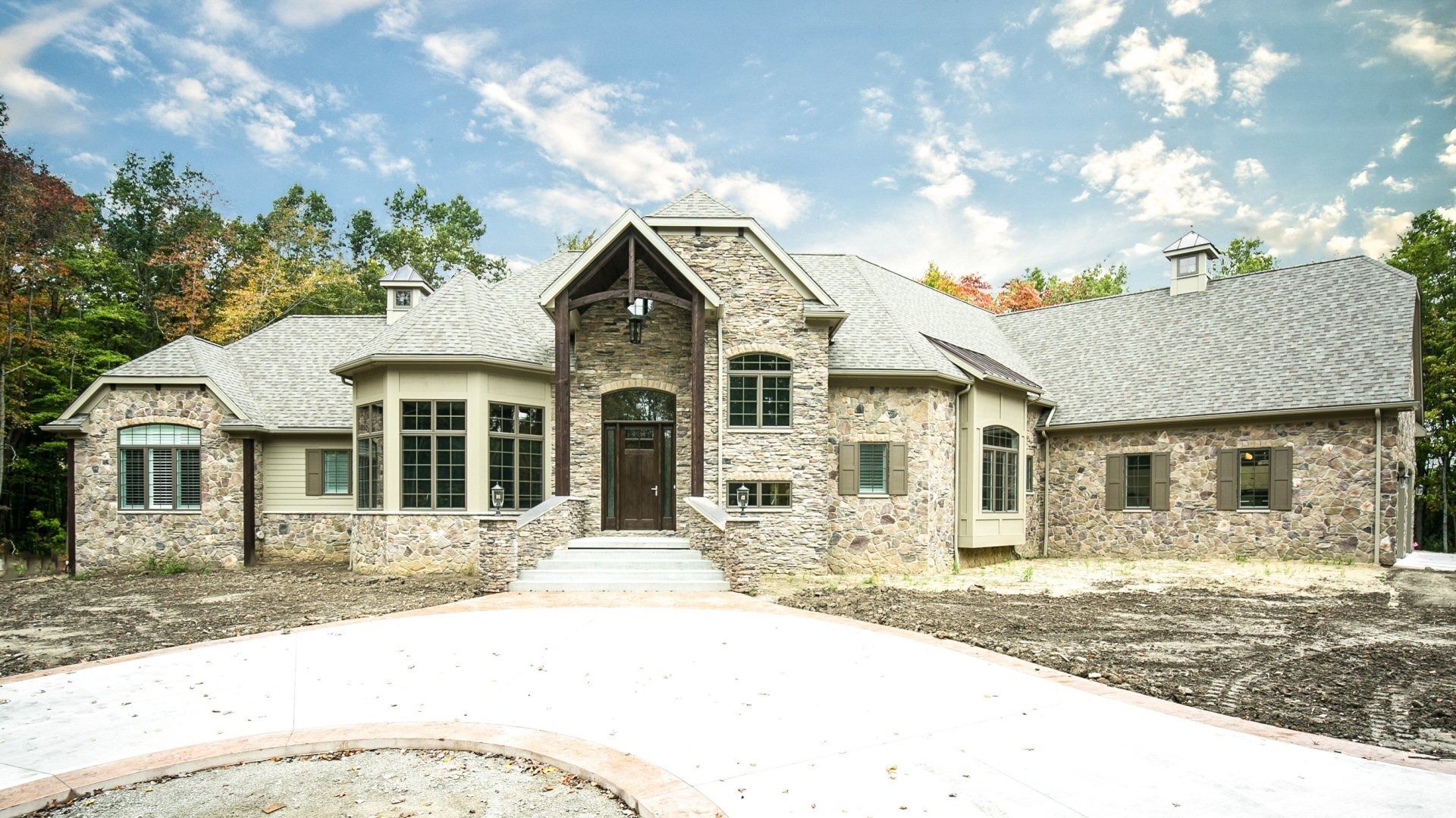 Stone-faced house with curved roof, arched entrance, and circular driveway. Cloudy sky, trees in the background.