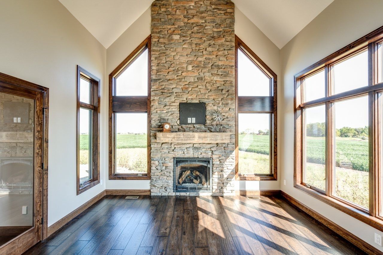 Empty living room with stone fireplace, large windows, and dark wood floors.