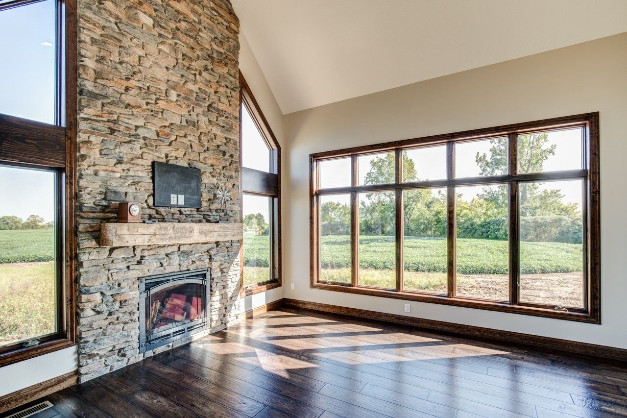 Living room with stone fireplace, large windows overlooking a field, and hardwood floor.