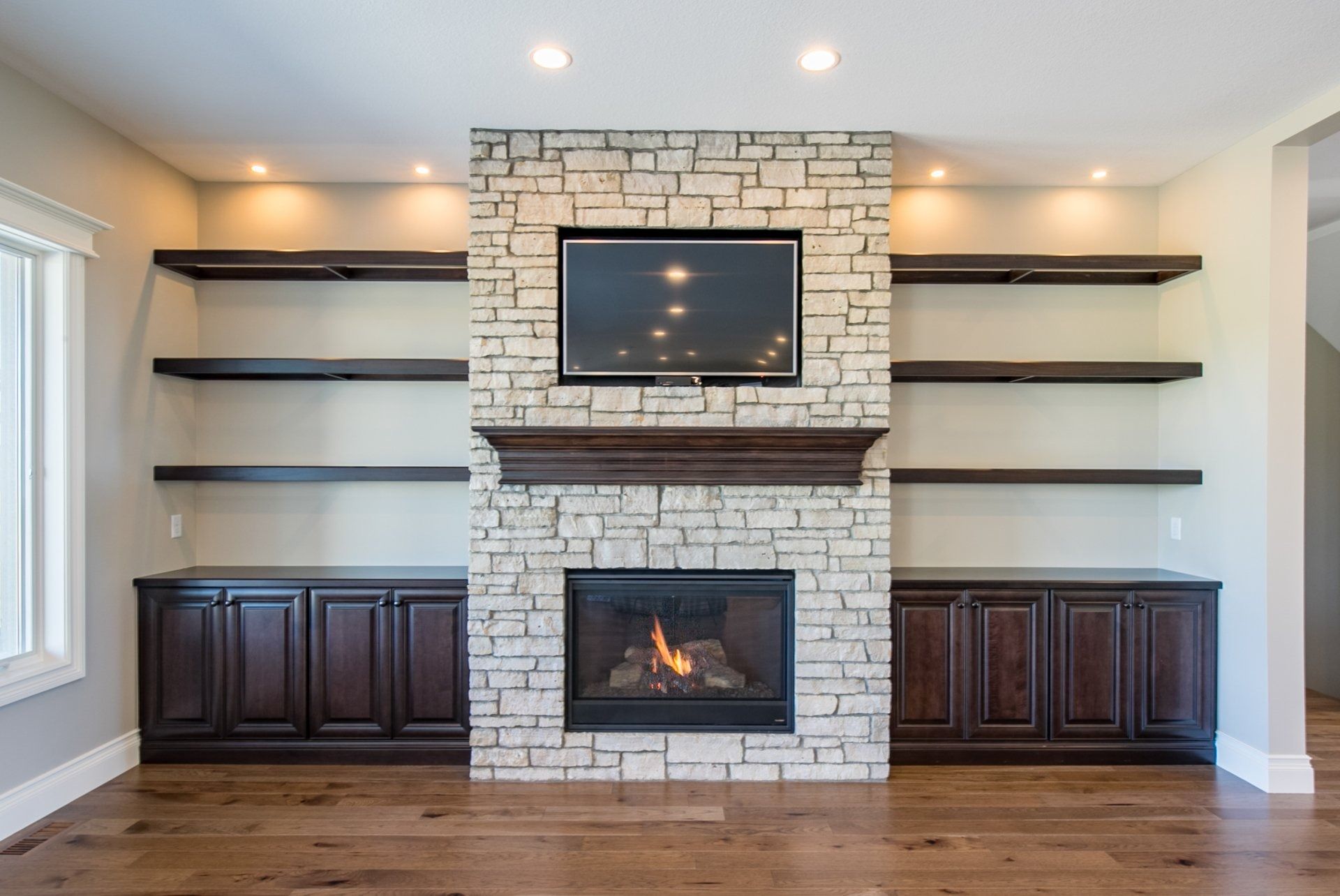 Living room with stone fireplace, built-in shelves, dark cabinets, TV, and recessed lighting.