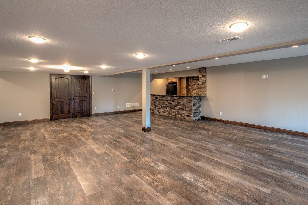 Empty basement with wood-look flooring, built-in bar with stone facade, dark wood double doors, and recessed lighting.
