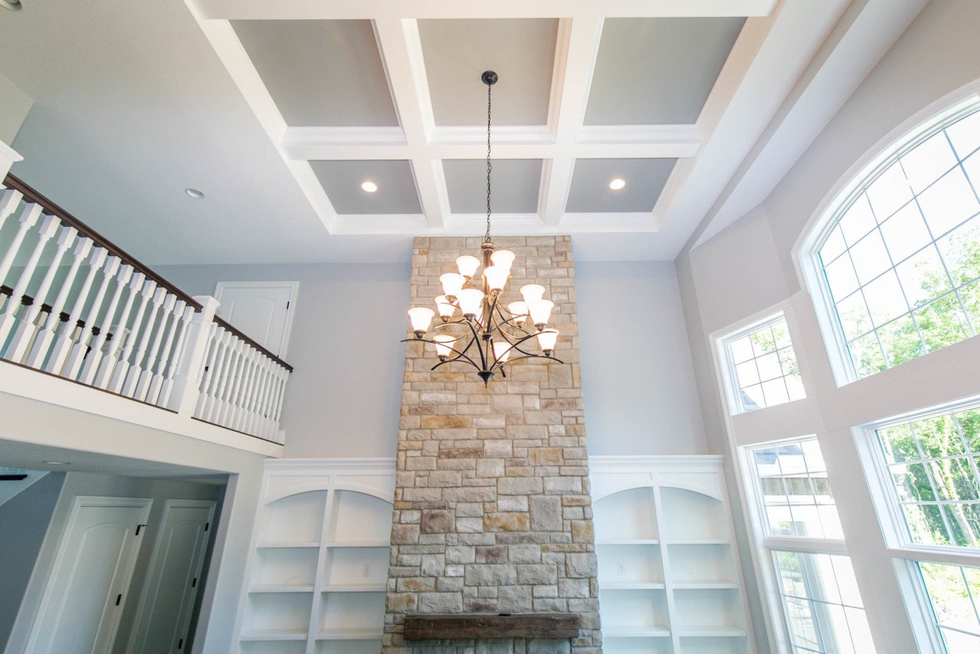High-ceilinged living room with stone fireplace, chandelier, built-in shelving, and large windows.