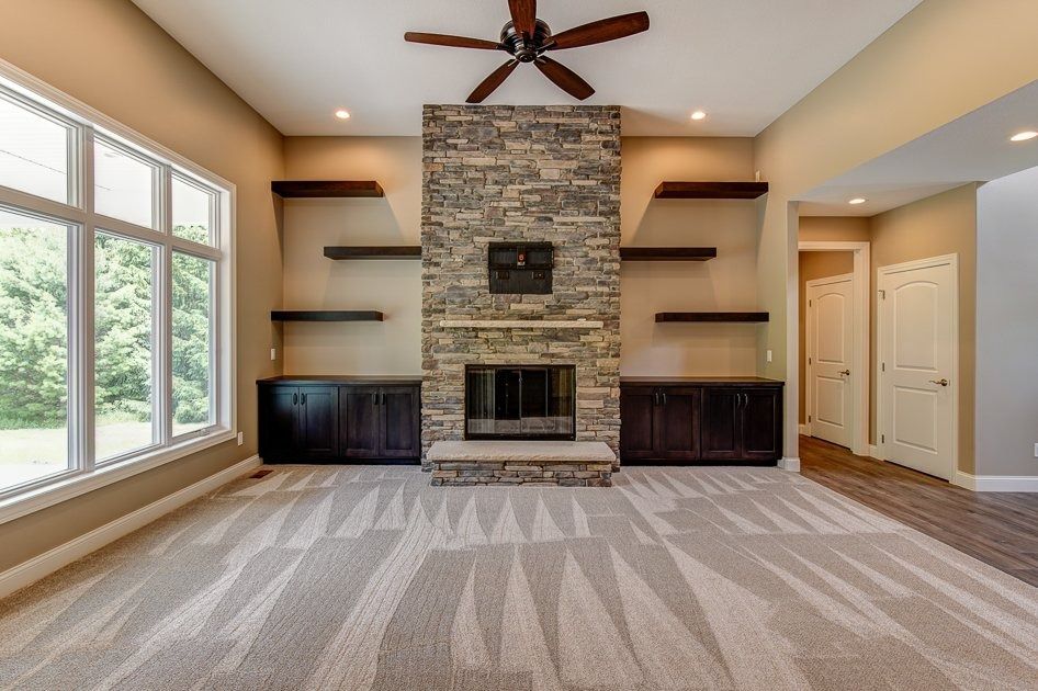 Living room with stone fireplace, built-in shelves, dark cabinets, patterned carpet, and large window.