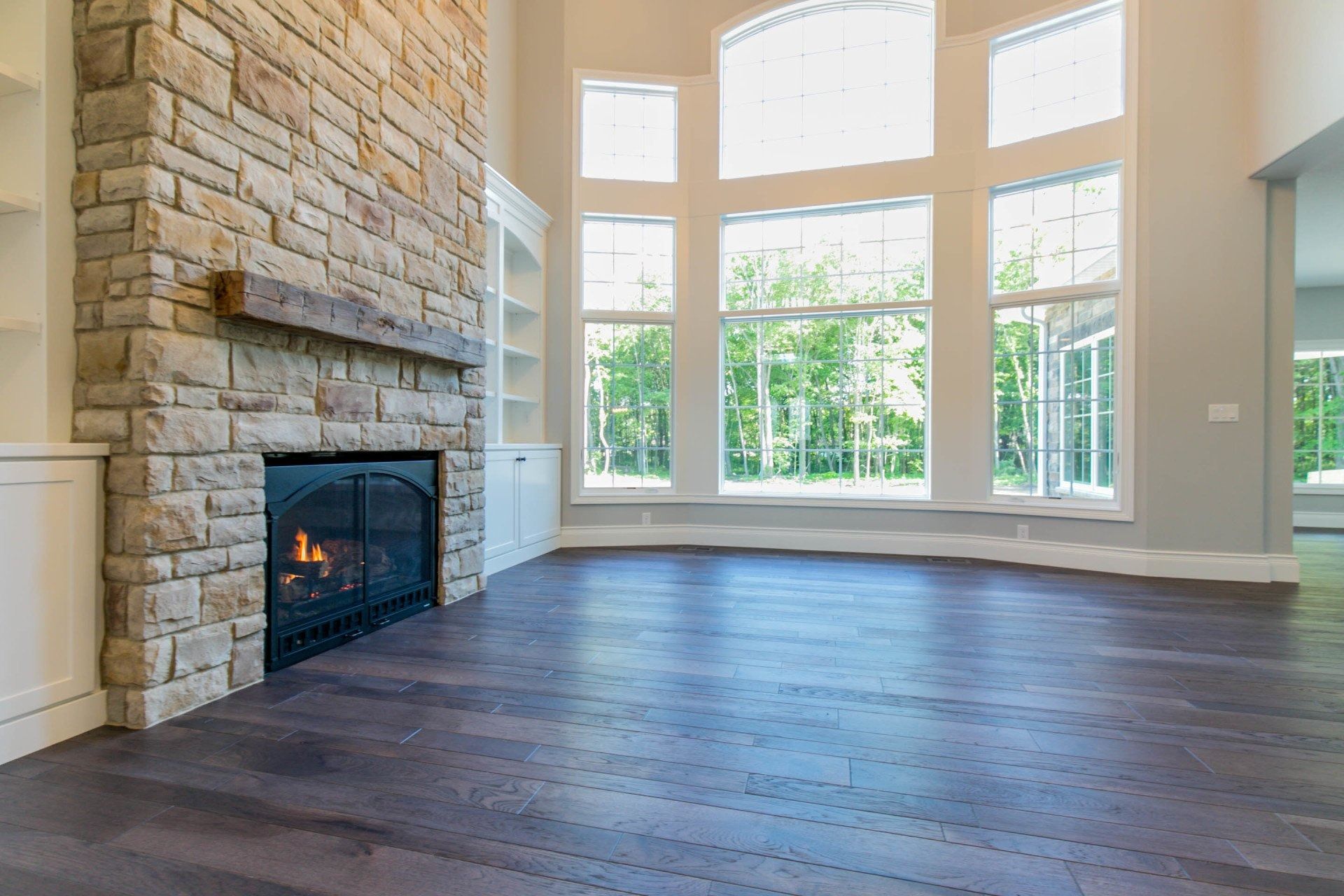 Stone fireplace in a bright living room with large windows overlooking trees and dark wood floor.