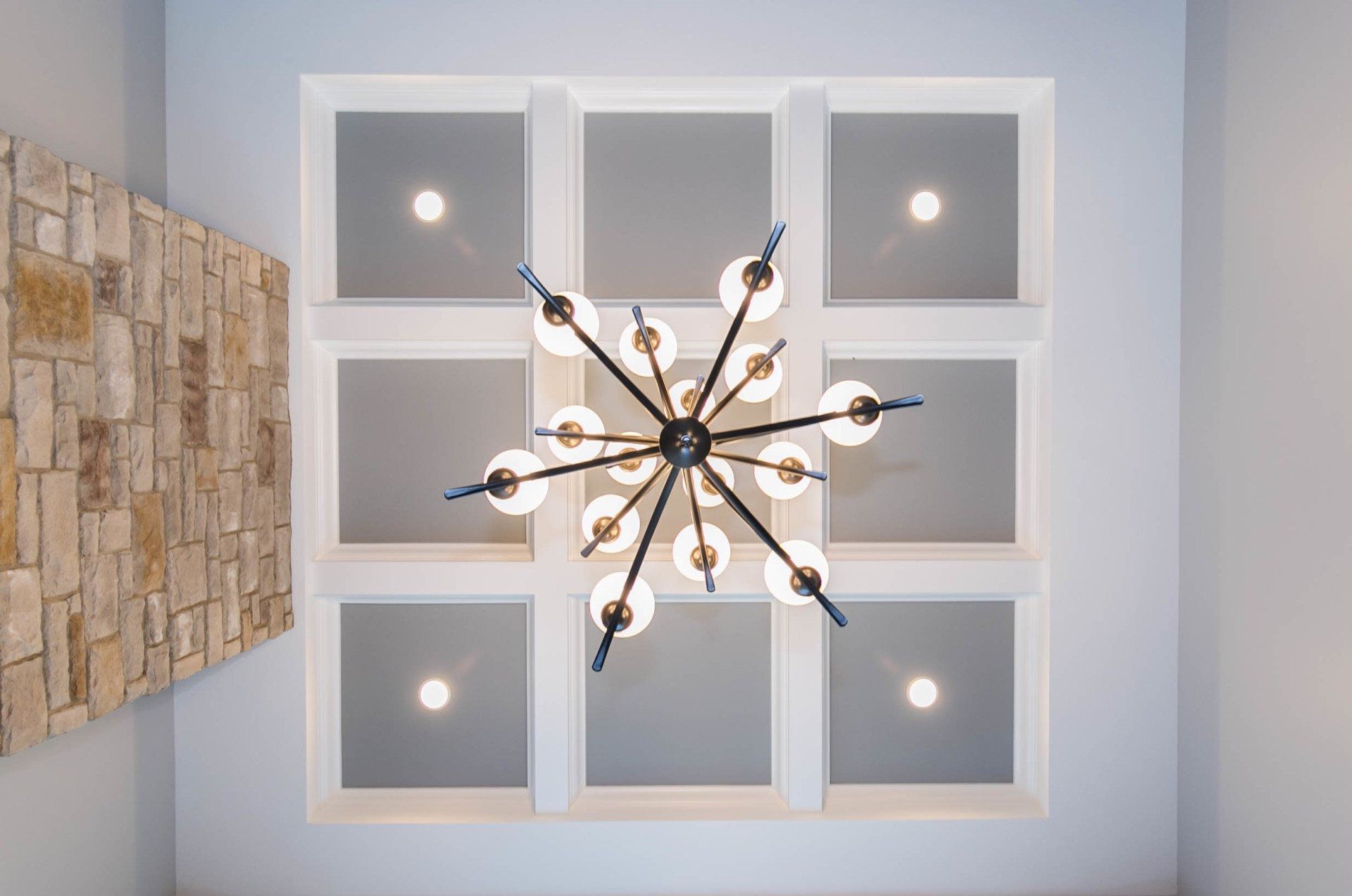 Chandelier hangs from a white grid ceiling with recessed lights, adjacent to a stone wall.